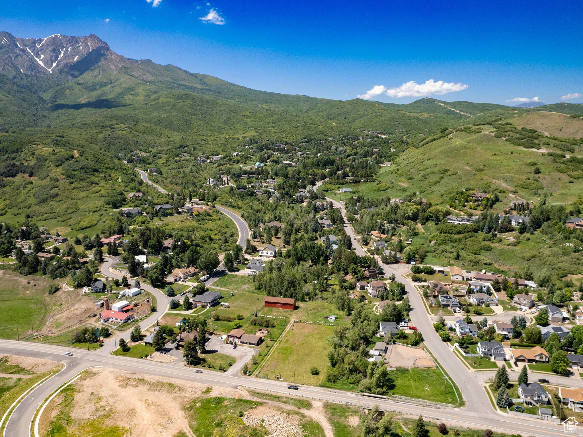 Expansive aerial showcasing a mountainous terrain with a snow-capped peak, abundant green foliage, and residential developments