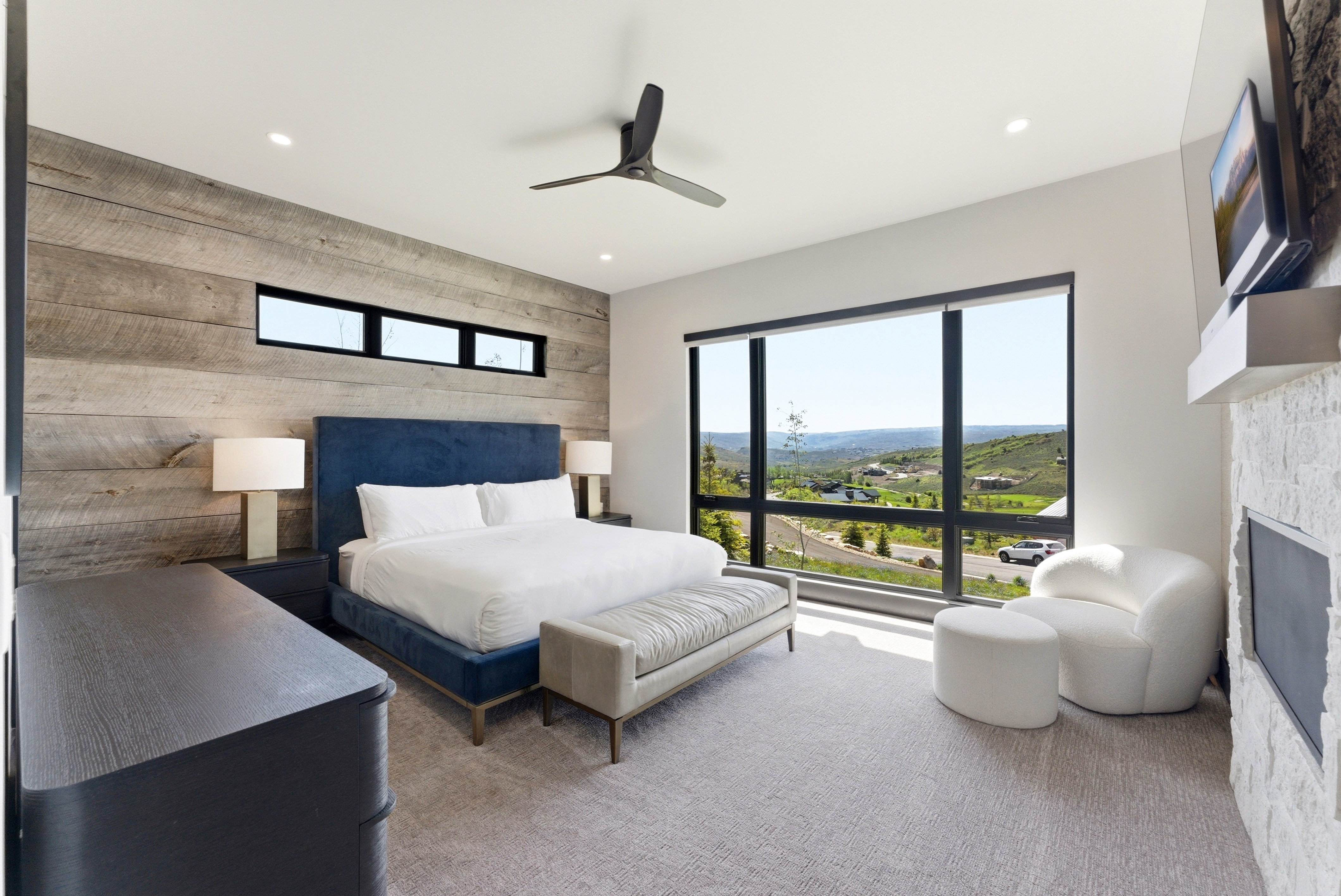 Bedroom featuring a full wood-paneled accent wall, a wide picture window, an integrated ceiling fan, recessed lighting, and a stone-clad fireplace