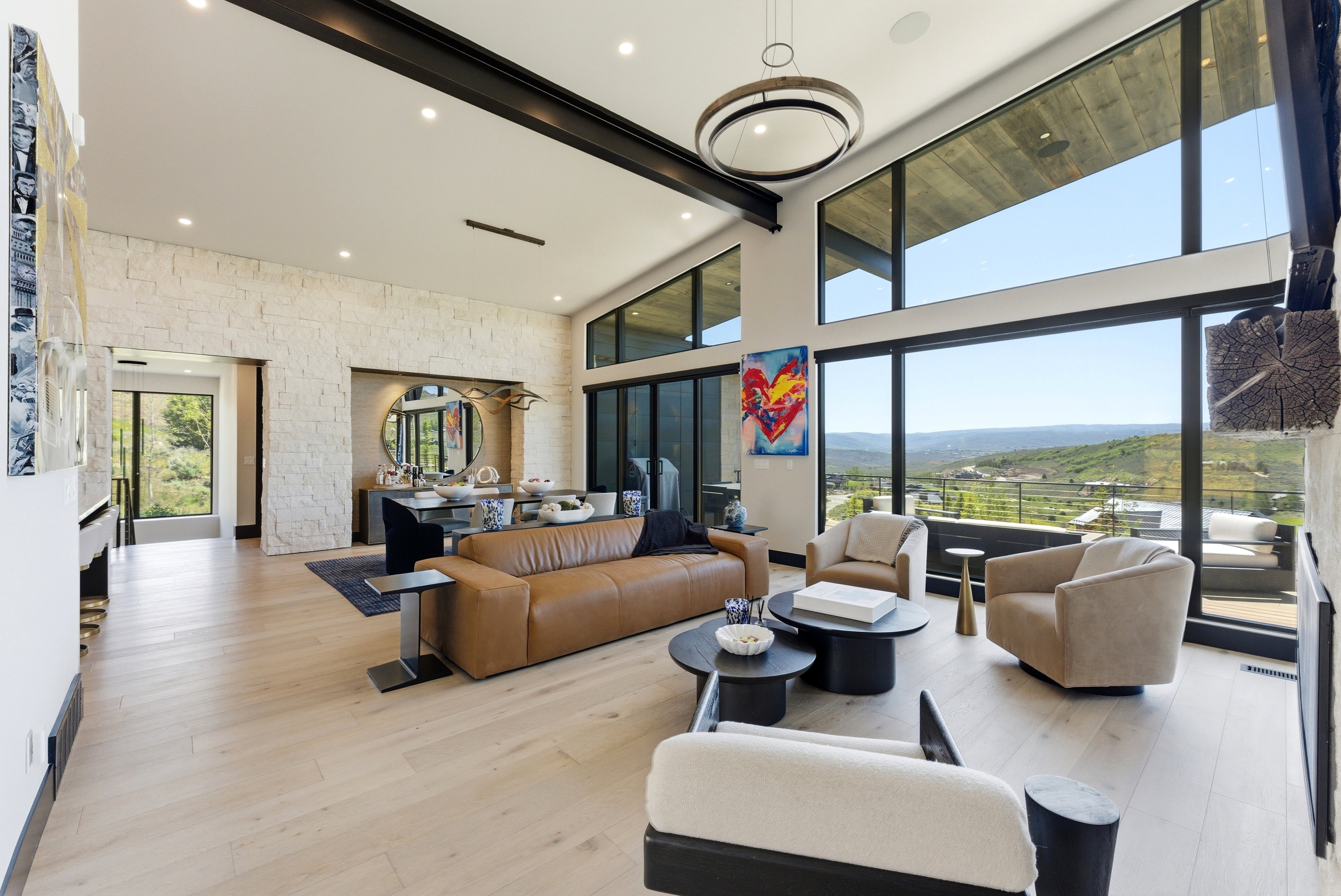 Spacious living area featuring expansive floor-to-ceiling windows, natural wood-finish flooring, a contemporary overhead lighting fixture, exposed ceiling beams, and a stone accent wall