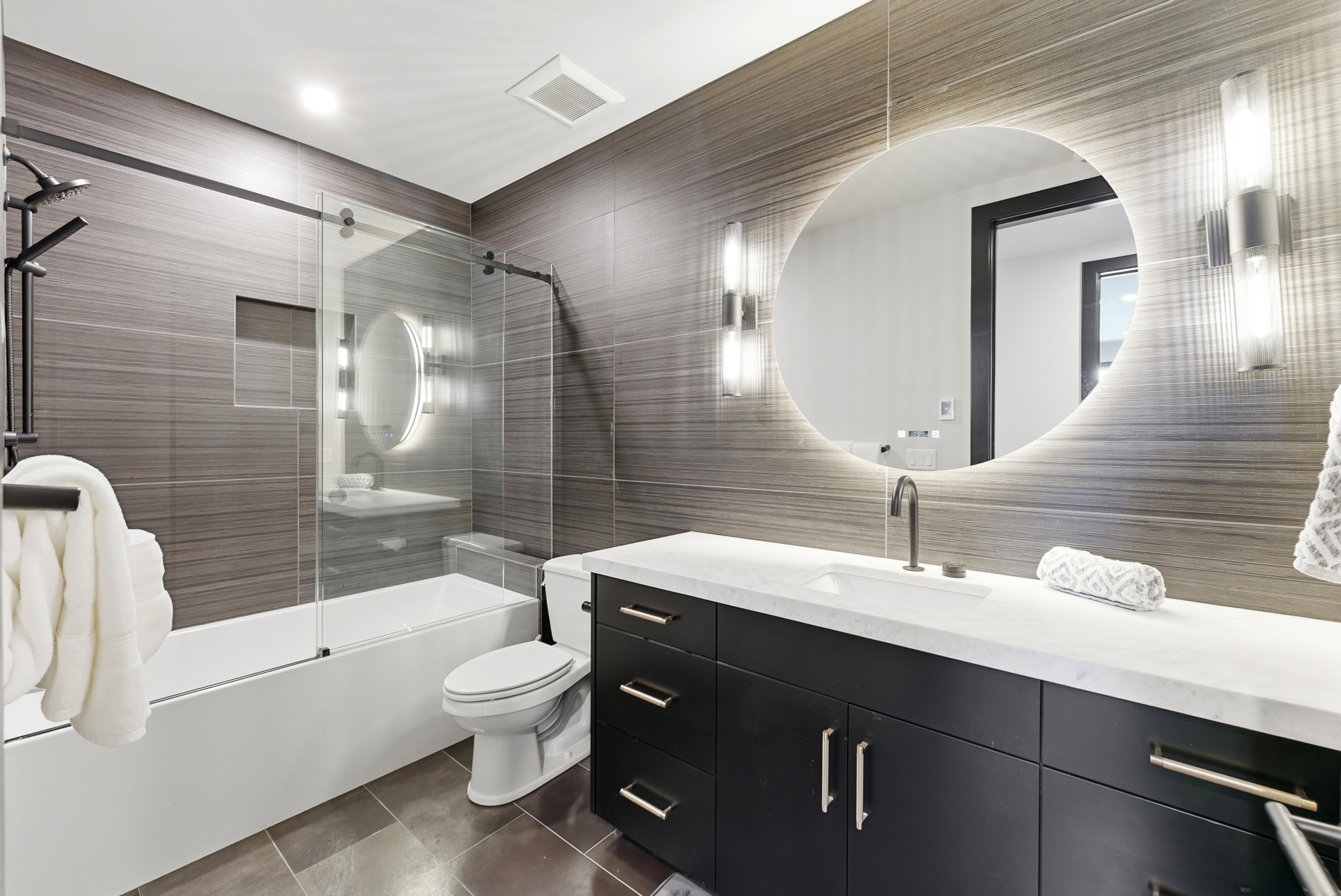 Bathroom featuring a deep soaking tub with glass shower enclosure, a dark wood-finish vanity with a white countertop, a circular back-lit mirror, and modern wall sconces