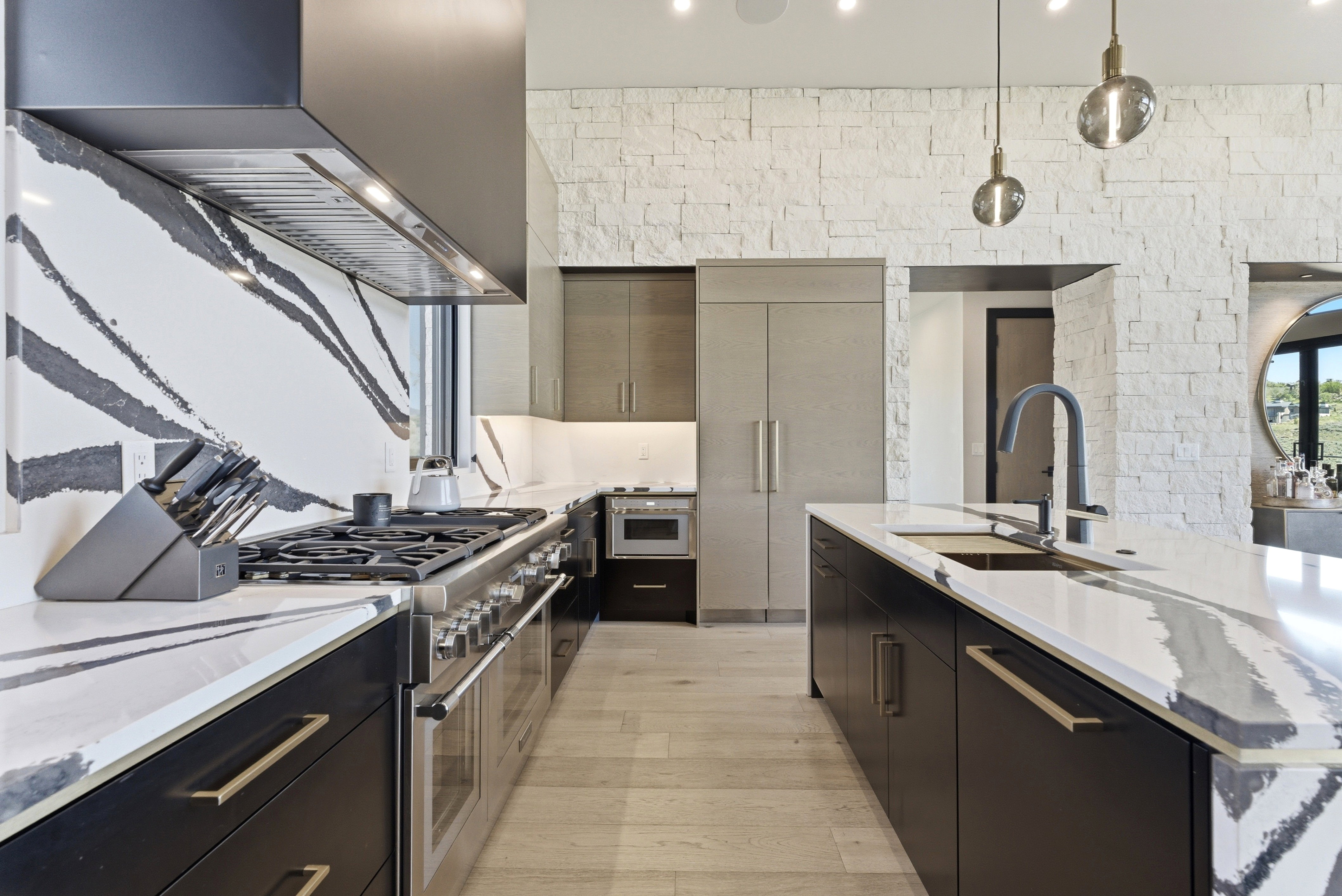 Gourmet kitchen featuring a professional-grade gas range, custom black cabinetry with gold hardware, a large kitchen island with integrated sink, and an accent wall of stacked stone veneer