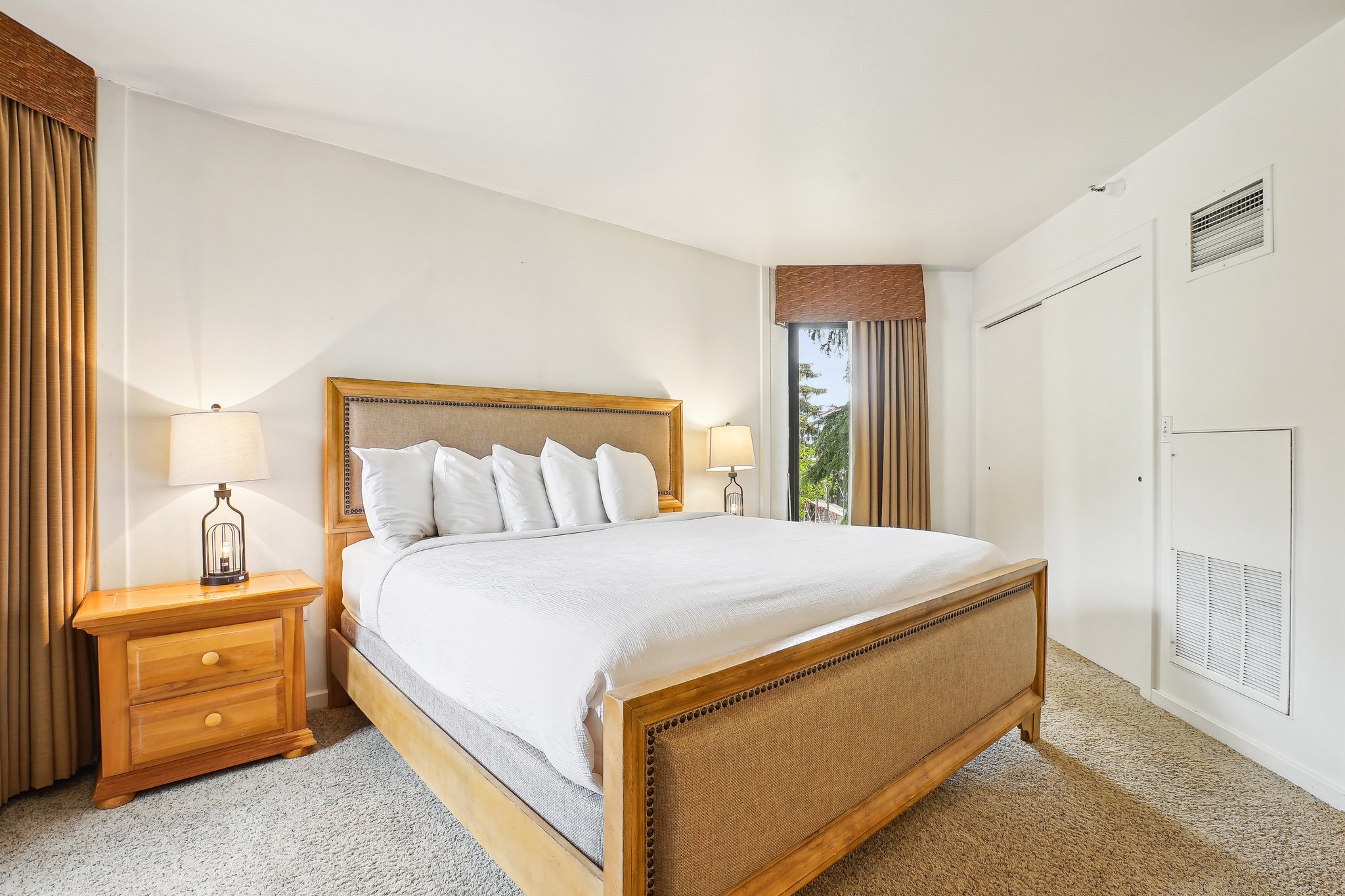 Carpeted bedroom featuring an upholstered bed frame, wooden nightstands, a large window with drapes, and a built-in sliding door closet