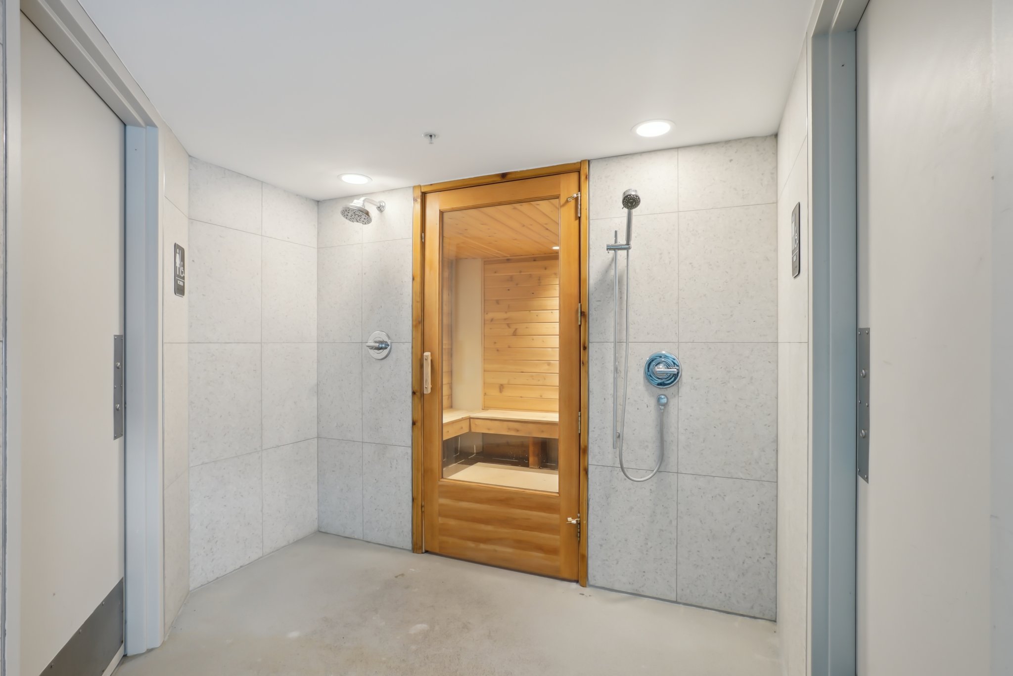 Indoor sauna with a clear-paneled wooden door, flanked by two shower areas featuring light-toned tile walls and chrome fixtures