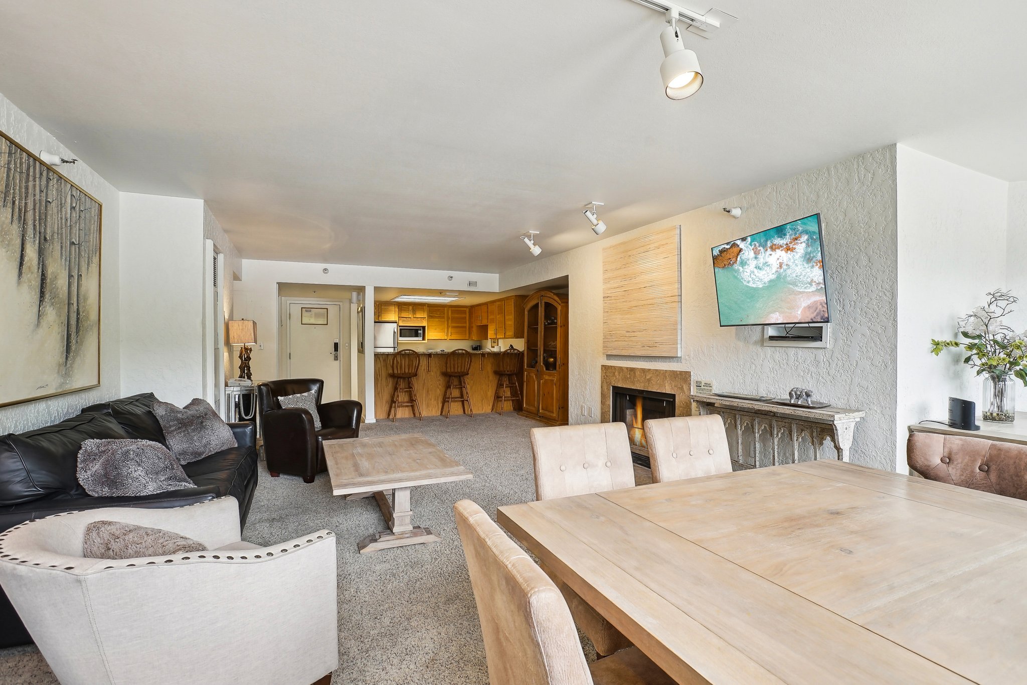 Open-concept living space featuring a dining area with a light wood-finish table, track lighting, a textured wall finish, and a built-in fireplace with a stone surround