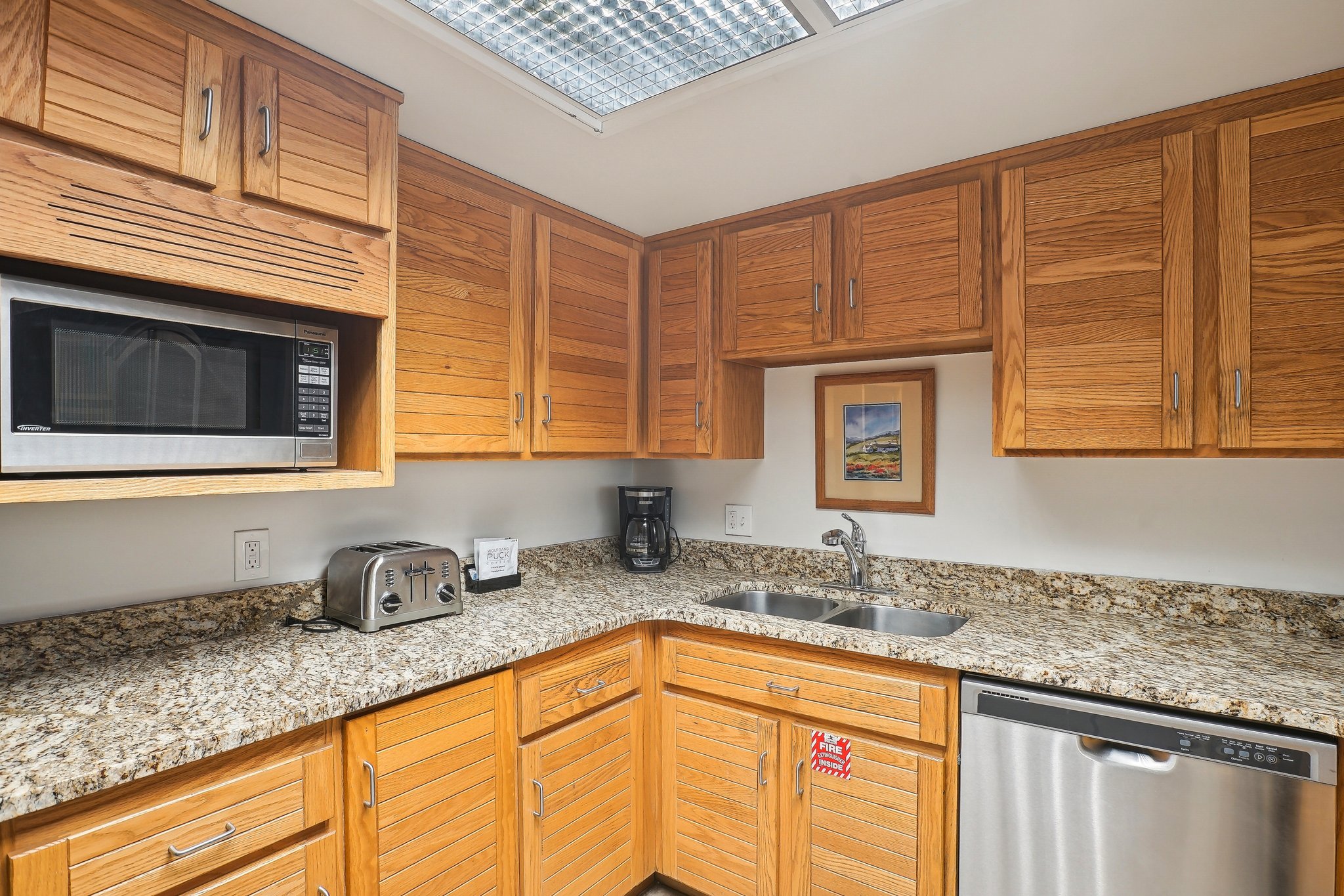 Corner kitchen featuring wood cabinetry, speckled countertops, a stainless steel dishwasher, a double basin sink, and a built-in microwave