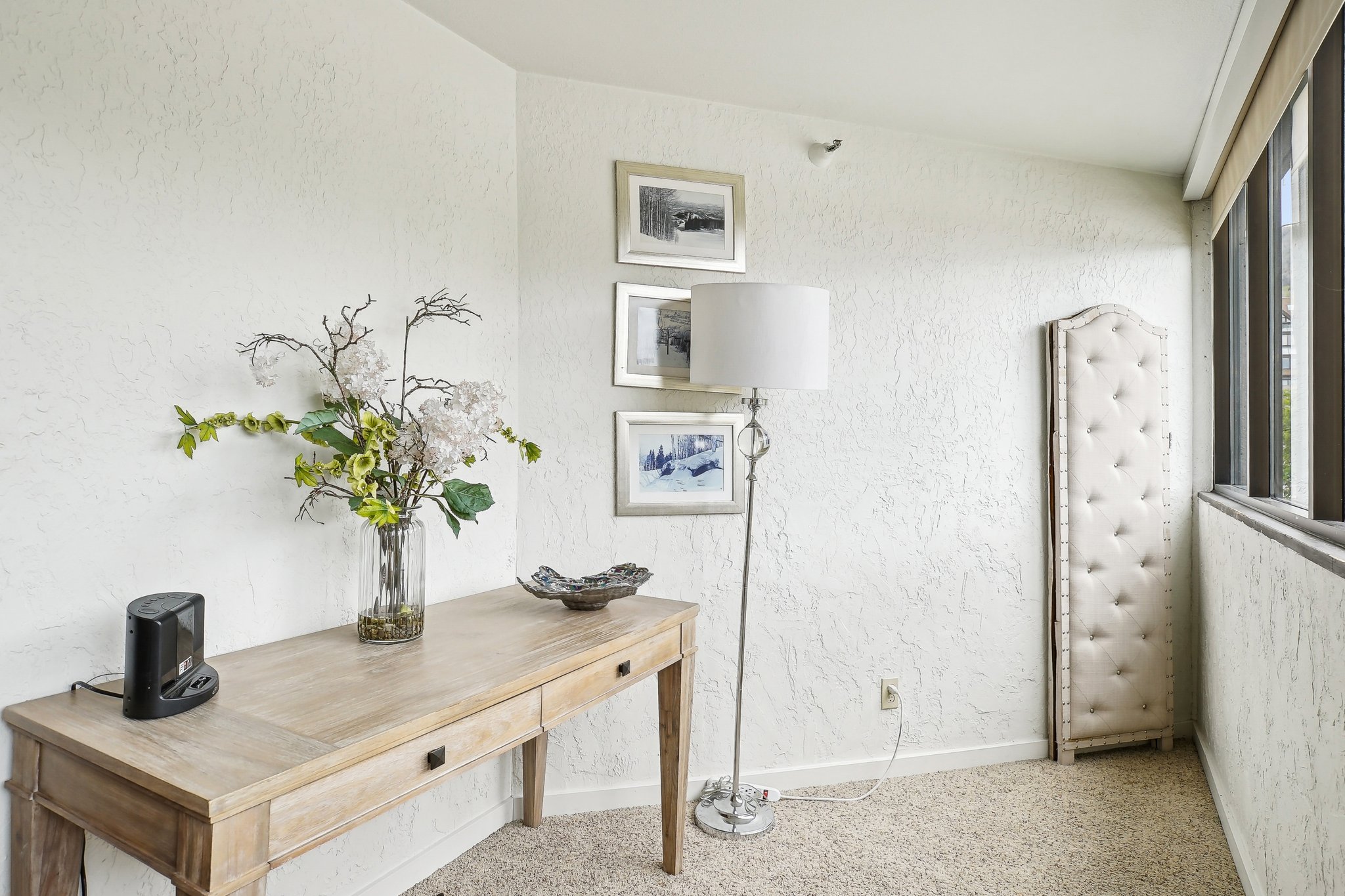Light-toned wood-finish console table with two drawers, textured white walls, light-colored carpet flooring, and a bank of windows providing natural light