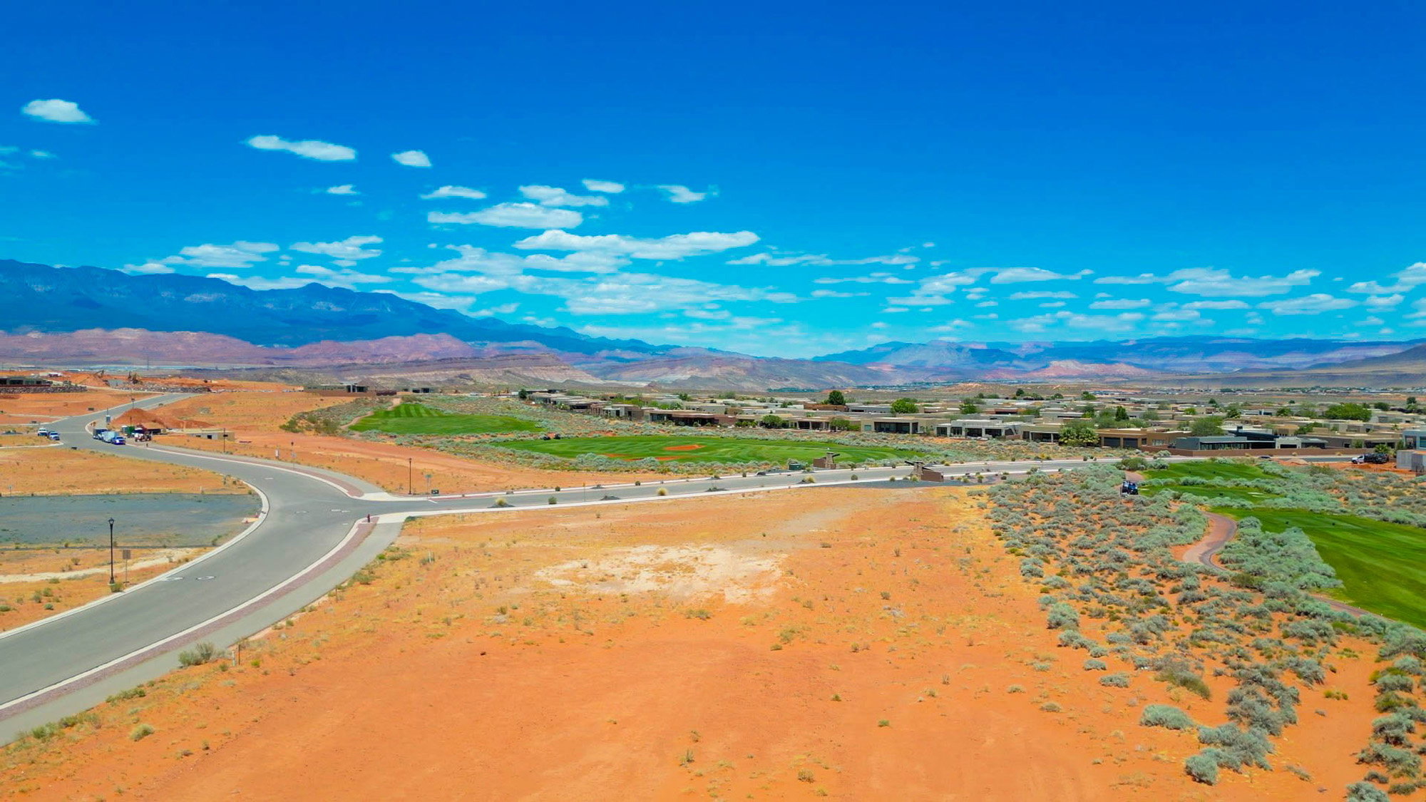 Expansive desert landscape featuring a paved road, scattered low-lying vegetation, a manicured golf course, and distant mountain ranges