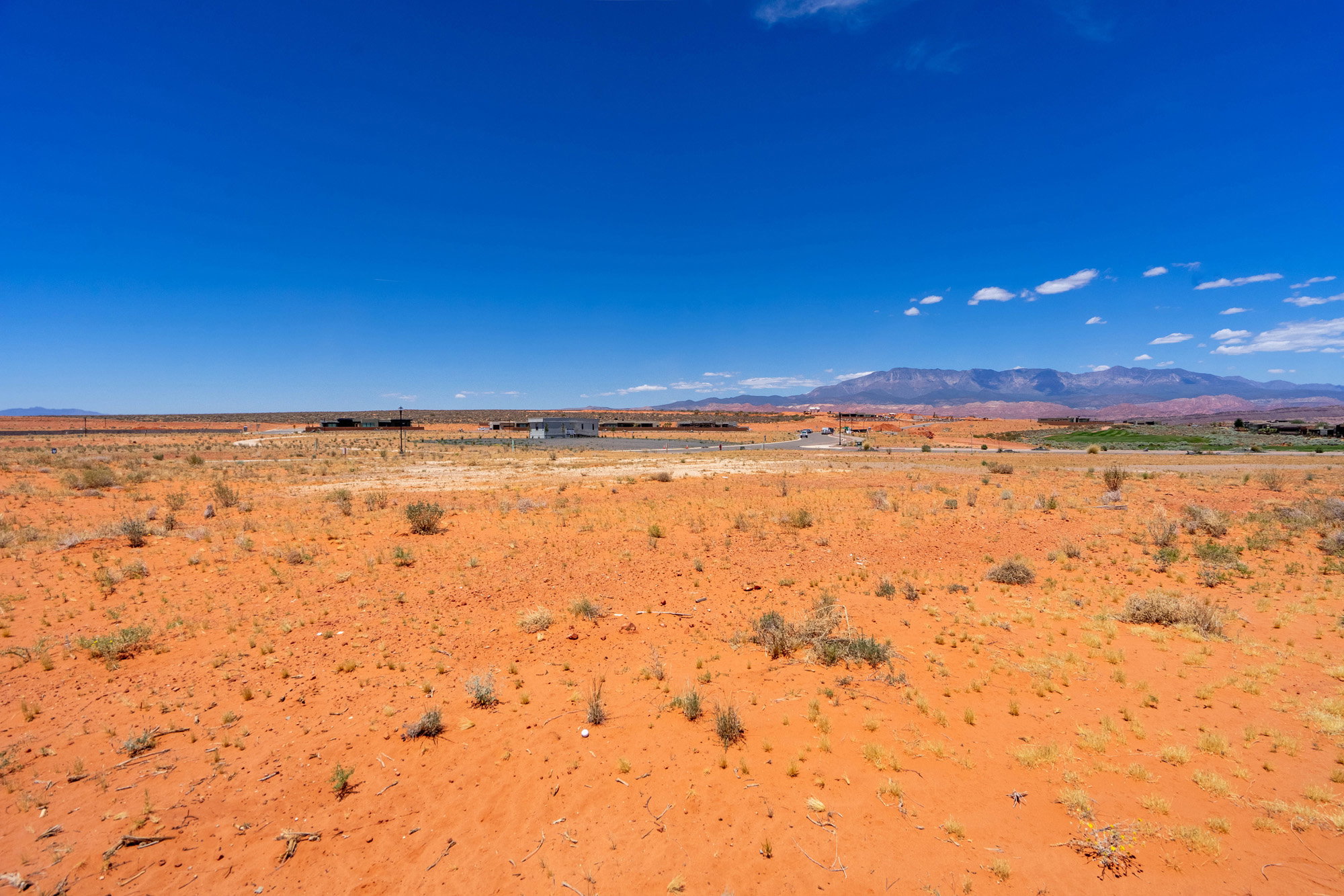 Expansive desert landscape featuring red earth, sparse vegetation, a distant mountain range, and a clear blue sky