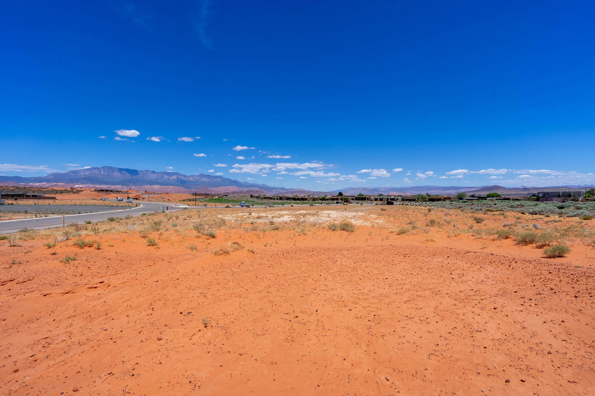 Expansive red sand terrain featuring scattered desert vegetation, a paved road, and distant mountain ranges