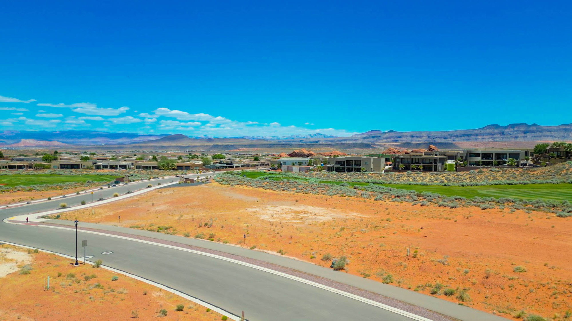 Expansive desert landscape featuring a paved roadway, undeveloped land, and distant structures with a backdrop of mountains