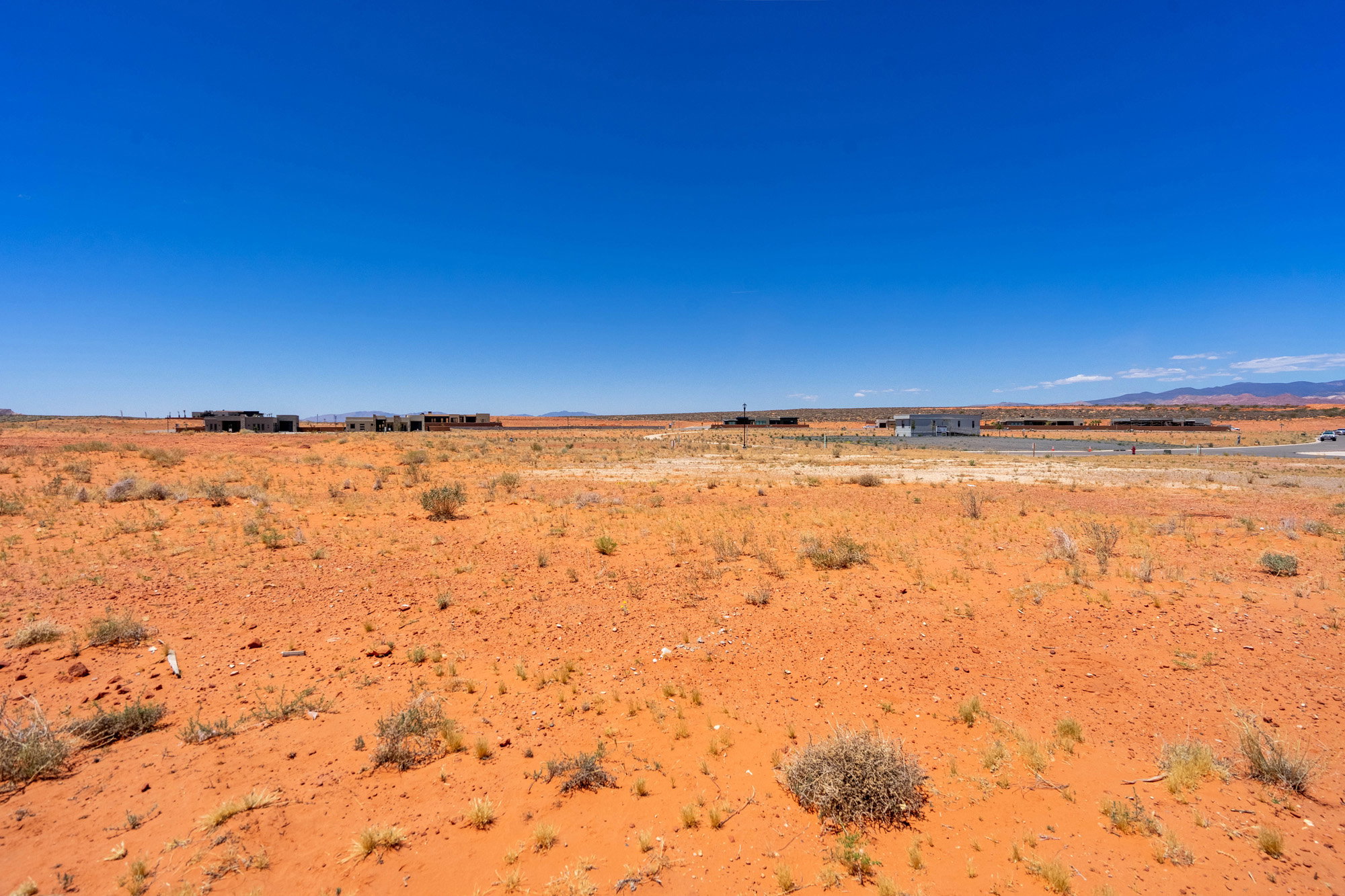 Expansive desert landscape featuring red earth, scattered low-lying vegetation, and distant structures under a clear blue sky
