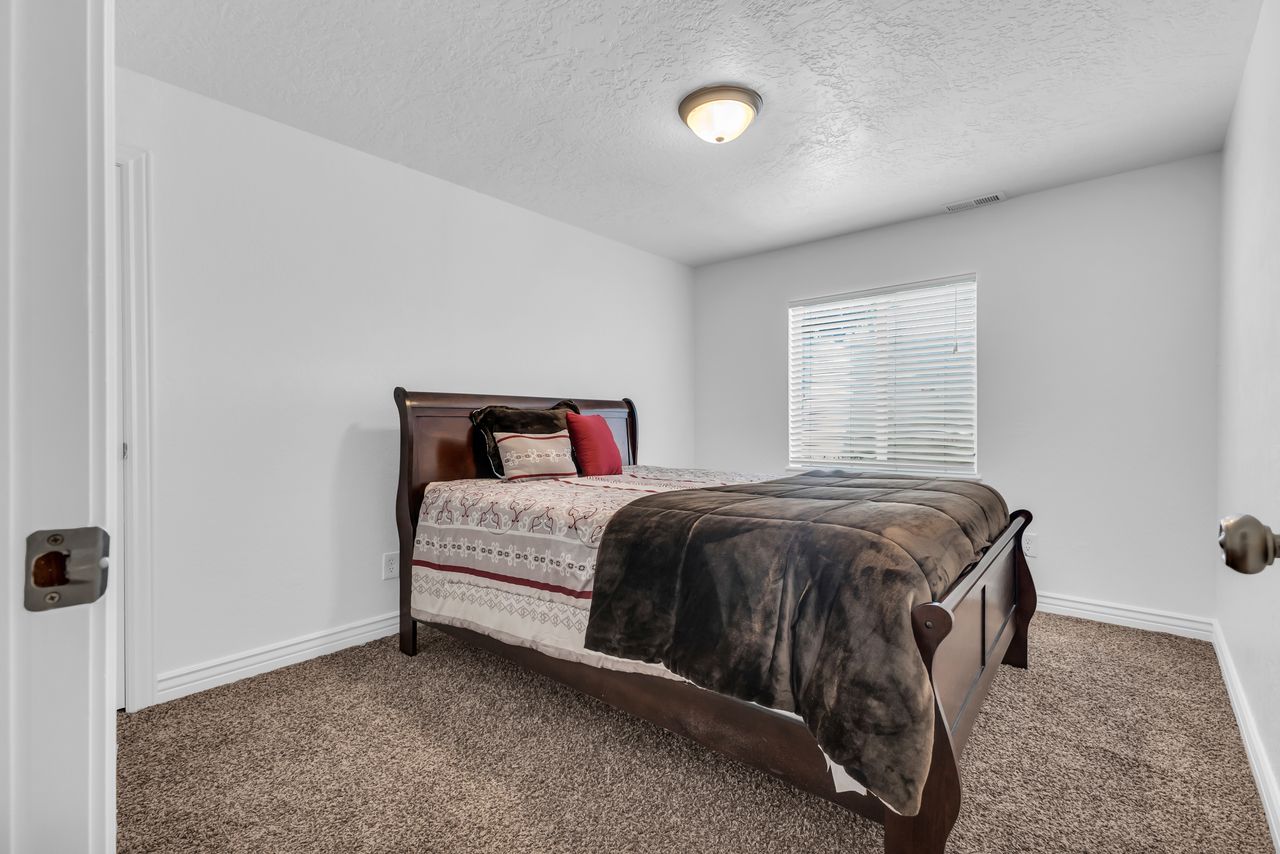 Bedroom featuring a window with blinds, a ceiling-mounted light fixture, white walls, and neutral-toned carpet flooring