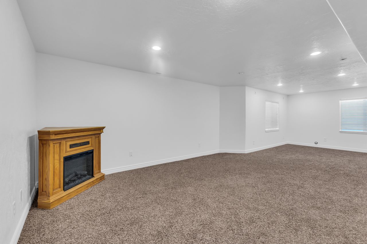 Spacious room featuring a corner-placed electric fireplace with a wood-finish mantel, neutral carpeting, recessed ceiling lighting, and multiple windows with blinds