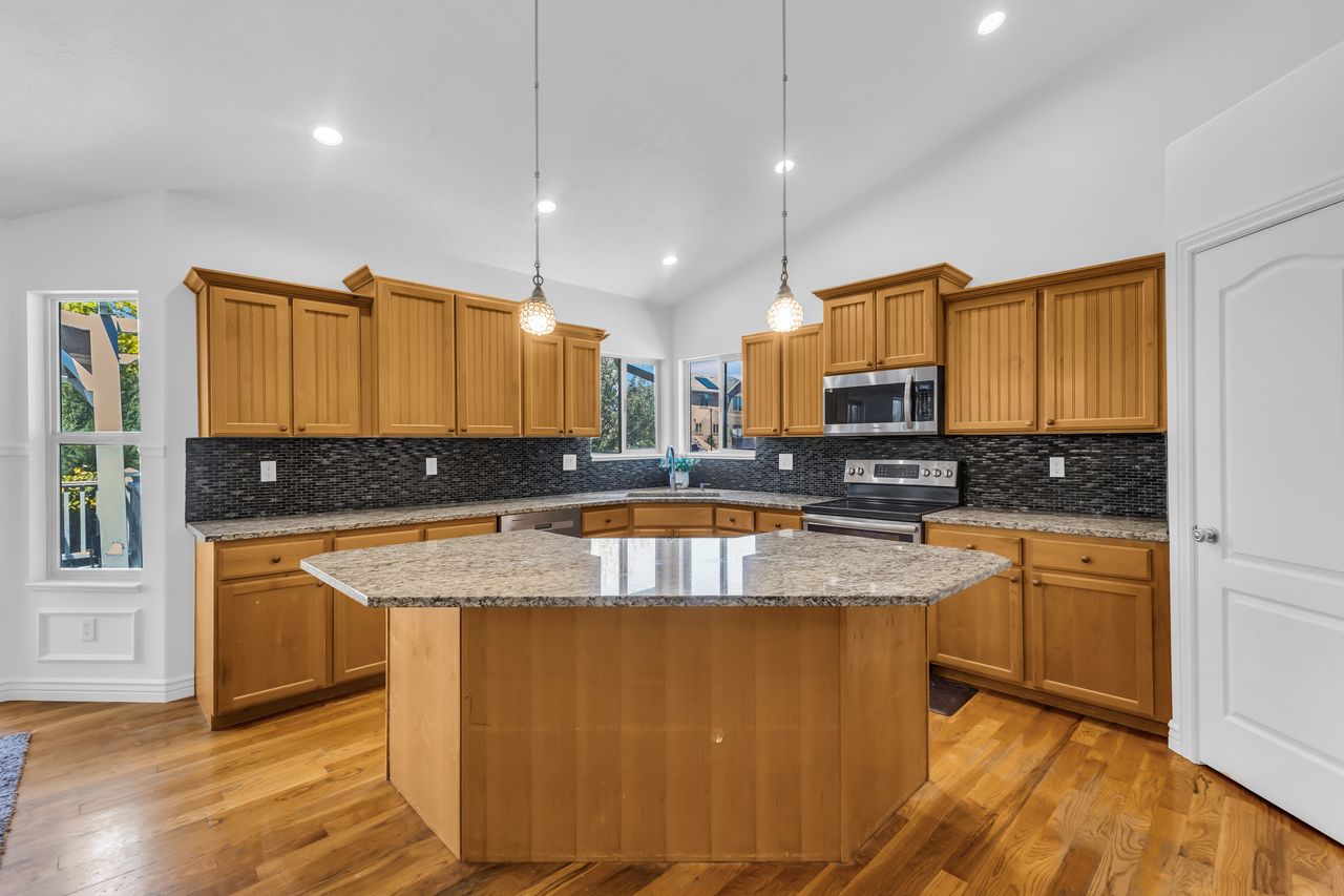 Kitchen featuring a multi-sided island with a light-toned countertop, wood-finish flooring, recessed lighting, pendant lights, and light wood cabinetry with dark tile backsplash