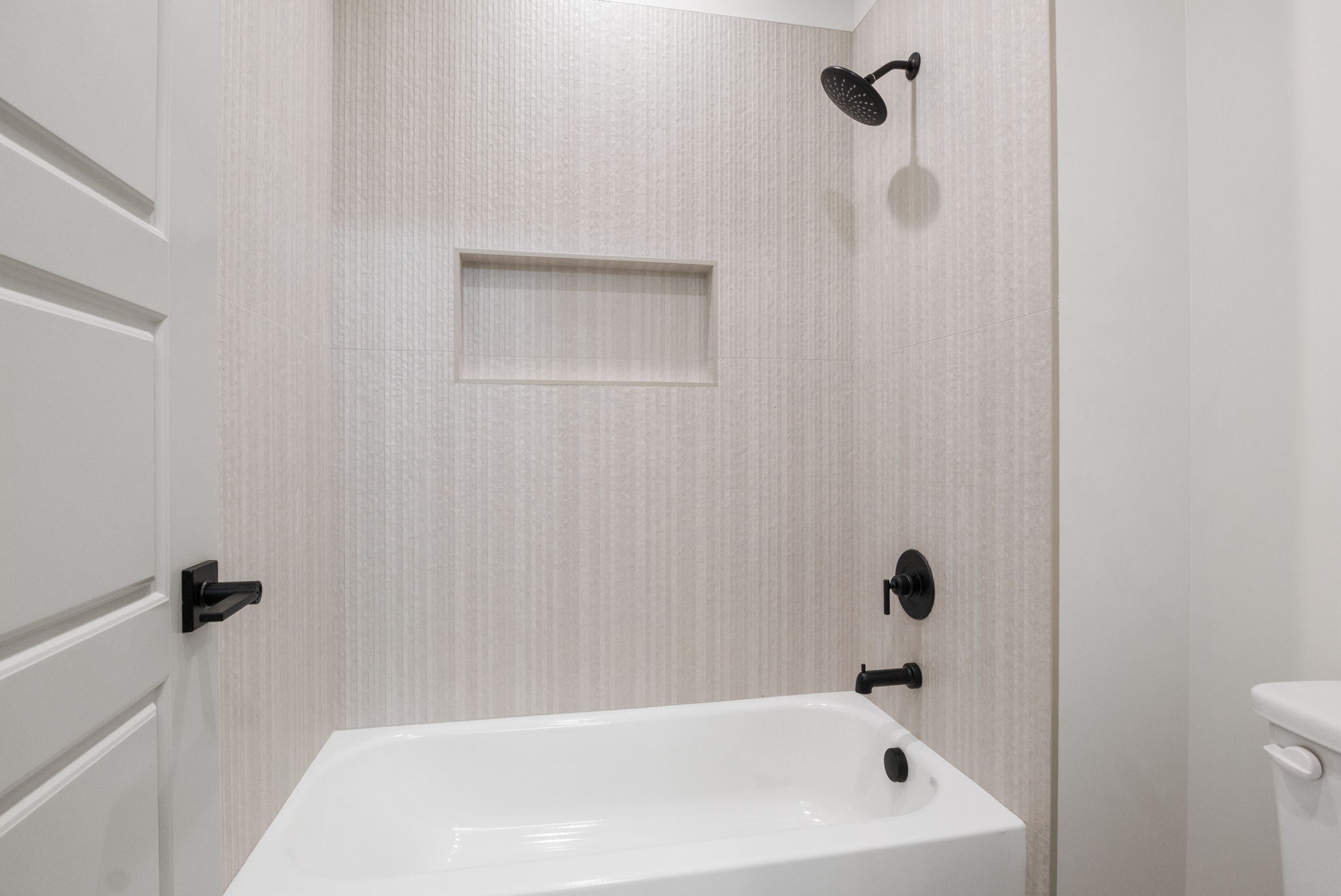 White bathtub with a matte black showerhead and fixtures, textured tiled shower surround, and a recessed shower niche