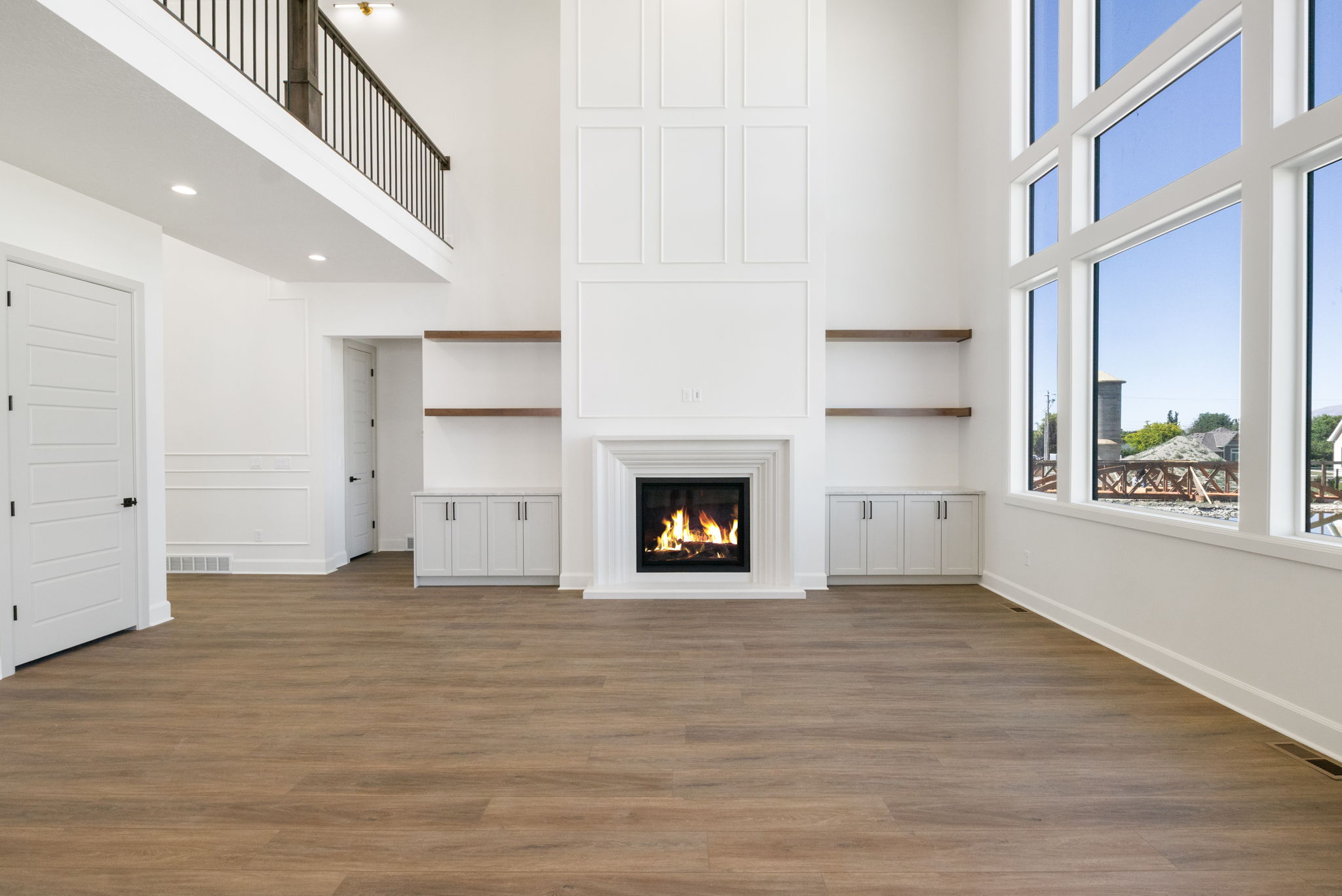 Two-story living area featuring a fireplace with a detailed mantel, built-in cabinetry with open shelving, an upper-level balcony with metal railing, and large multi-pane windows
