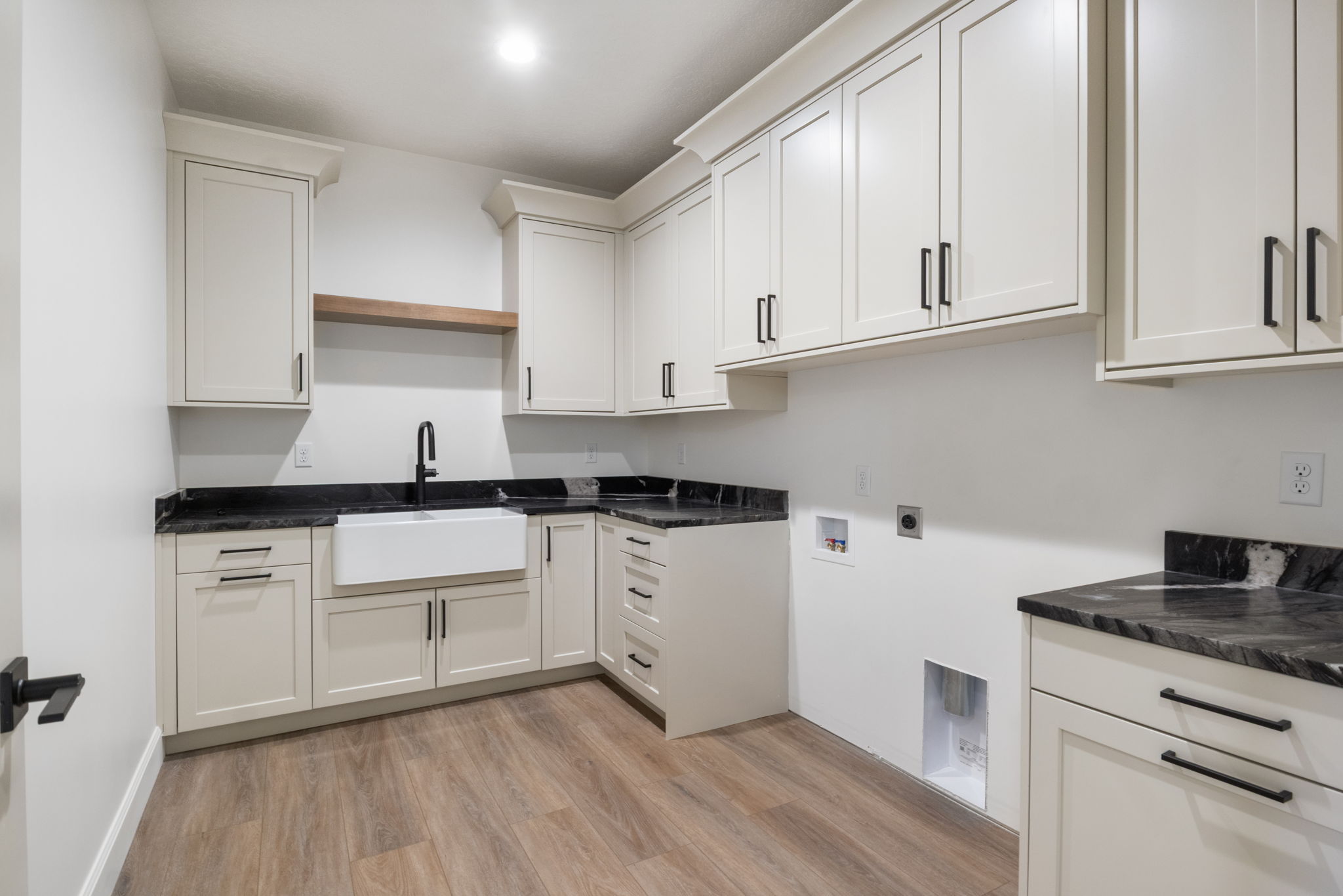 Utility room featuring extensive cabinetry with black hardware, dark countertops, a farmhouse-style sink with a matte black faucet, and wood-finish flooring