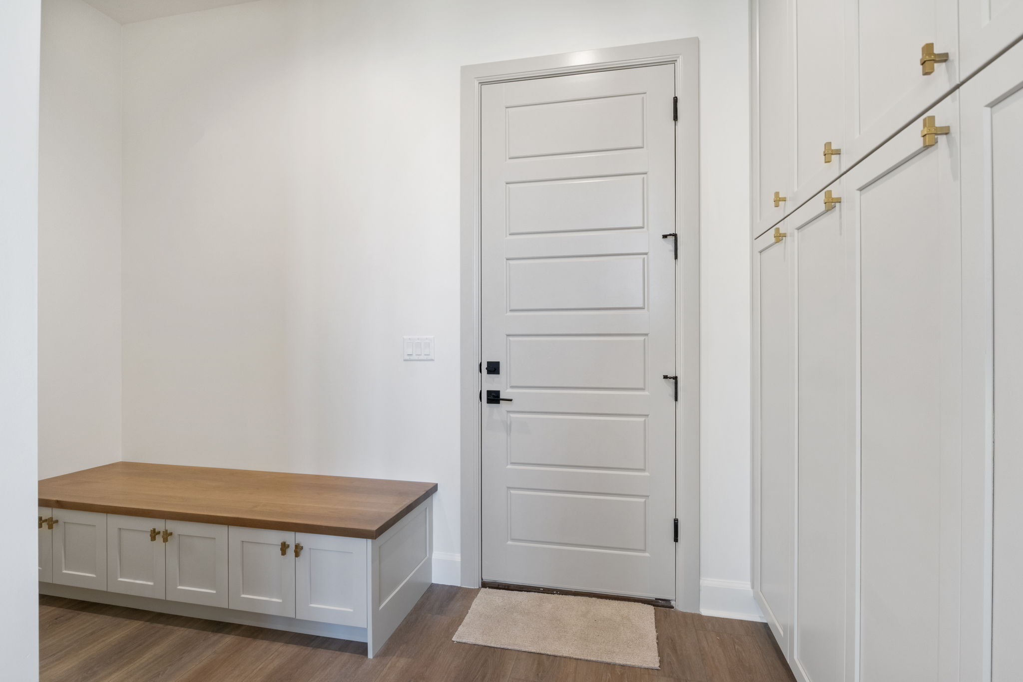 Entryway featuring wood-finish flooring, a built-in bench with a light wood countertop, white cabinetry with gold-finish hardware, a paneled door with black hardware, and floor-to-ceiling white storage cabinets with gold-finish hardware