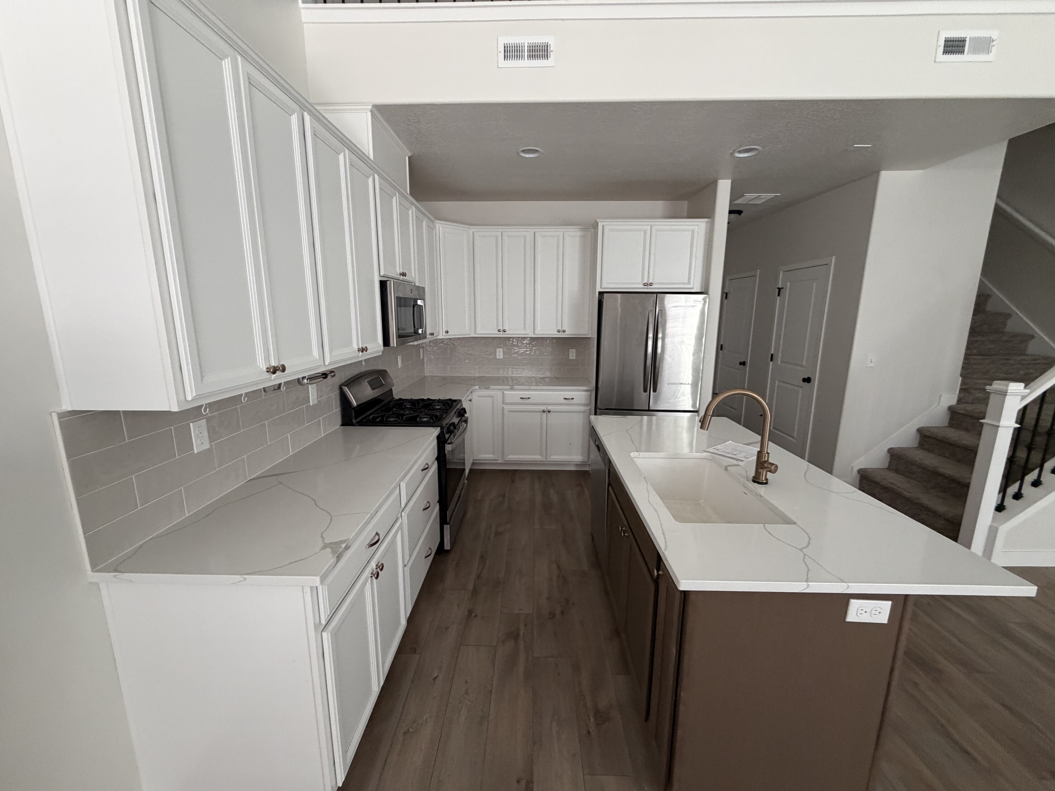 Contemporary kitchen featuring white shaker cabinetry, light-toned countertops with subtle veining, stainless steel appliances, a subway tile backsplash, and a central island with an integrated sink and bronze-finish faucet