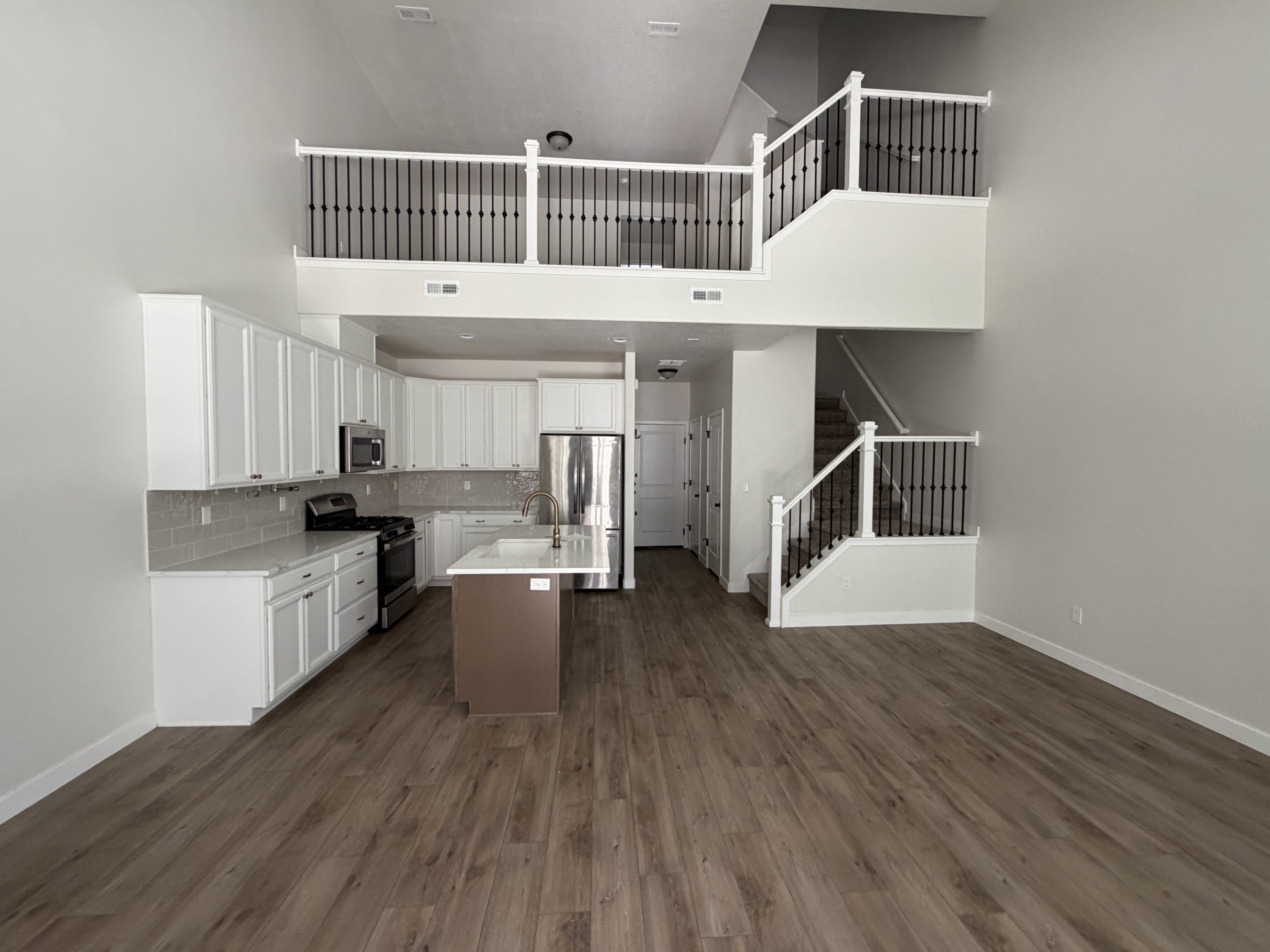 Open-concept living area featuring high ceilings, wood-finish flooring, a contemporary kitchen with white cabinetry, stainless steel appliances, and a central island, and a staircase leading to an upper-level loft