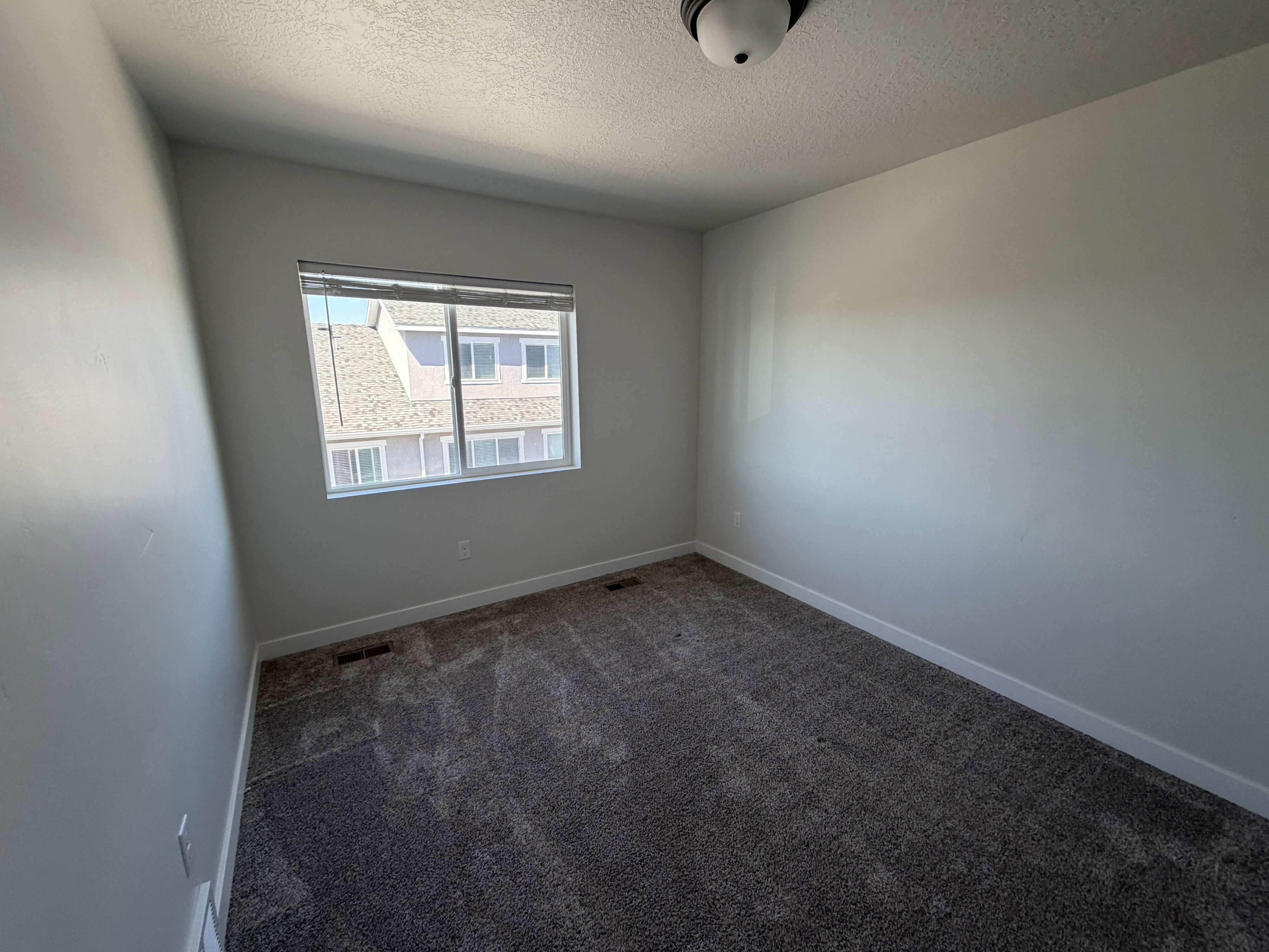 Room featuring a window with blinds, a ceiling-mounted light fixture, light gray walls, white baseboards, and dark gray patterned carpet