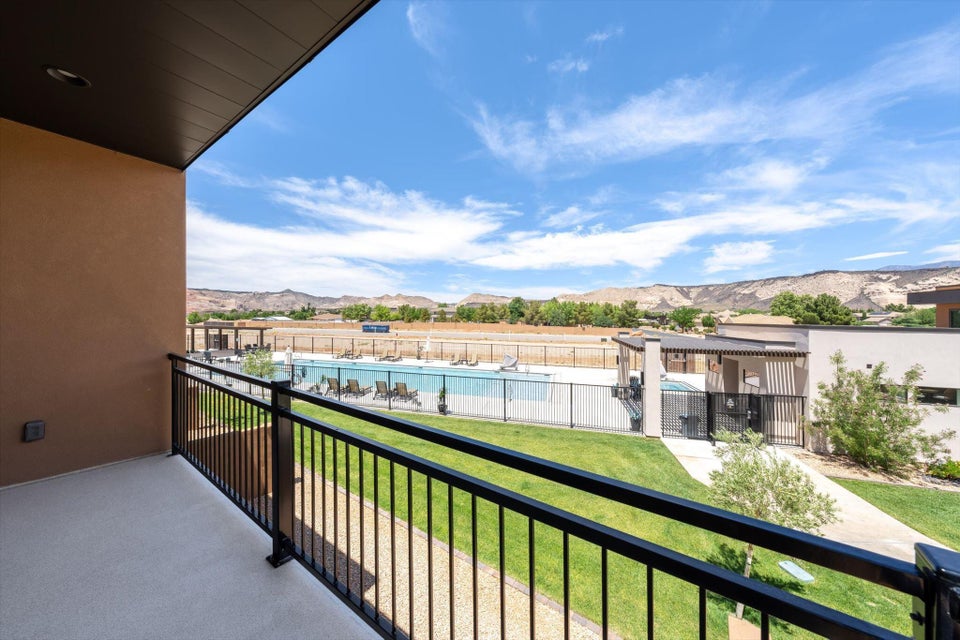 Balcony featuring a black metal railing and a textured exterior wall, overlooking a community pool with an adjacent lounge area, a manicured lawn, and distant natural terrain