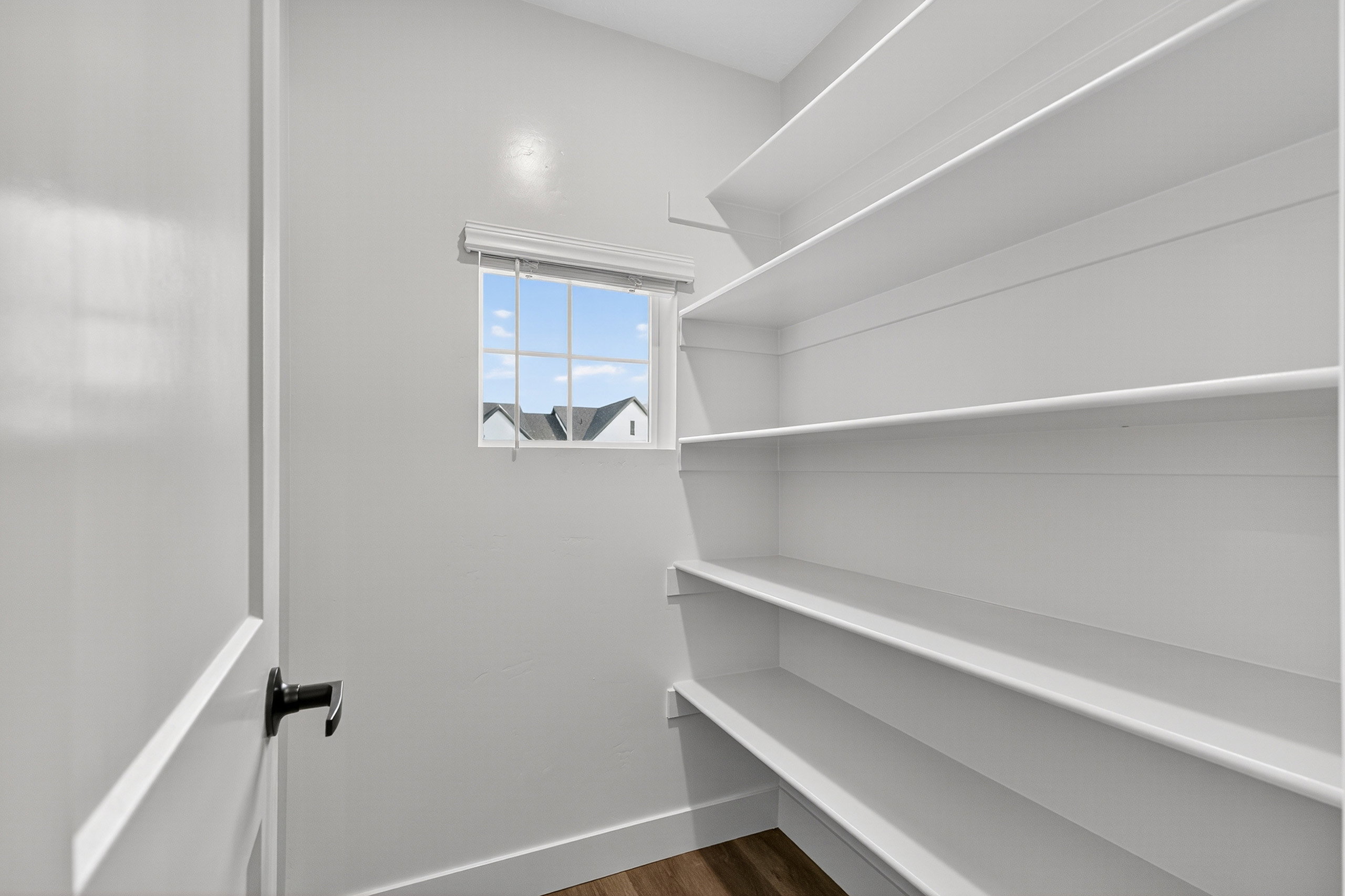 Spacious pantry featuring extensive built-in shelving, natural light from a window, wood-finish flooring, and a modern door with a black handle