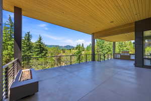 Expansive covered outdoor living area featuring a wood-finish ceiling, built-in outdoor kitchen, concrete flooring, and a linear fire pit with a metal railing