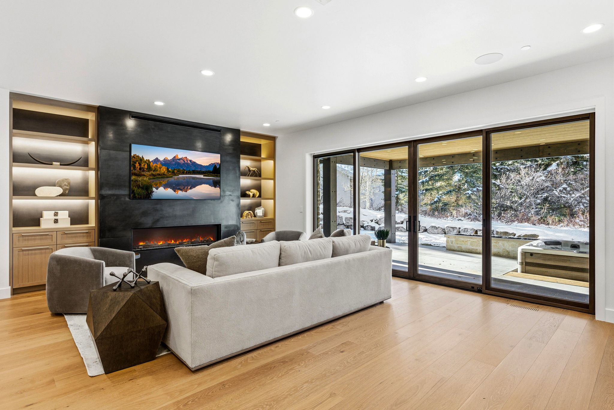 Spacious living area featuring light wood-finish flooring, a wall-mounted television above a linear fireplace, custom built-in shelving with integrated lighting, and expansive glass sliding doors opening to an outdoor patio with a hot tub