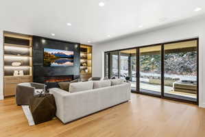 Spacious living area featuring light wood-finish flooring, a wall-mounted television above a linear fireplace, custom built-in shelving with integrated lighting, and expansive glass sliding doors opening to an outdoor patio with a hot tub