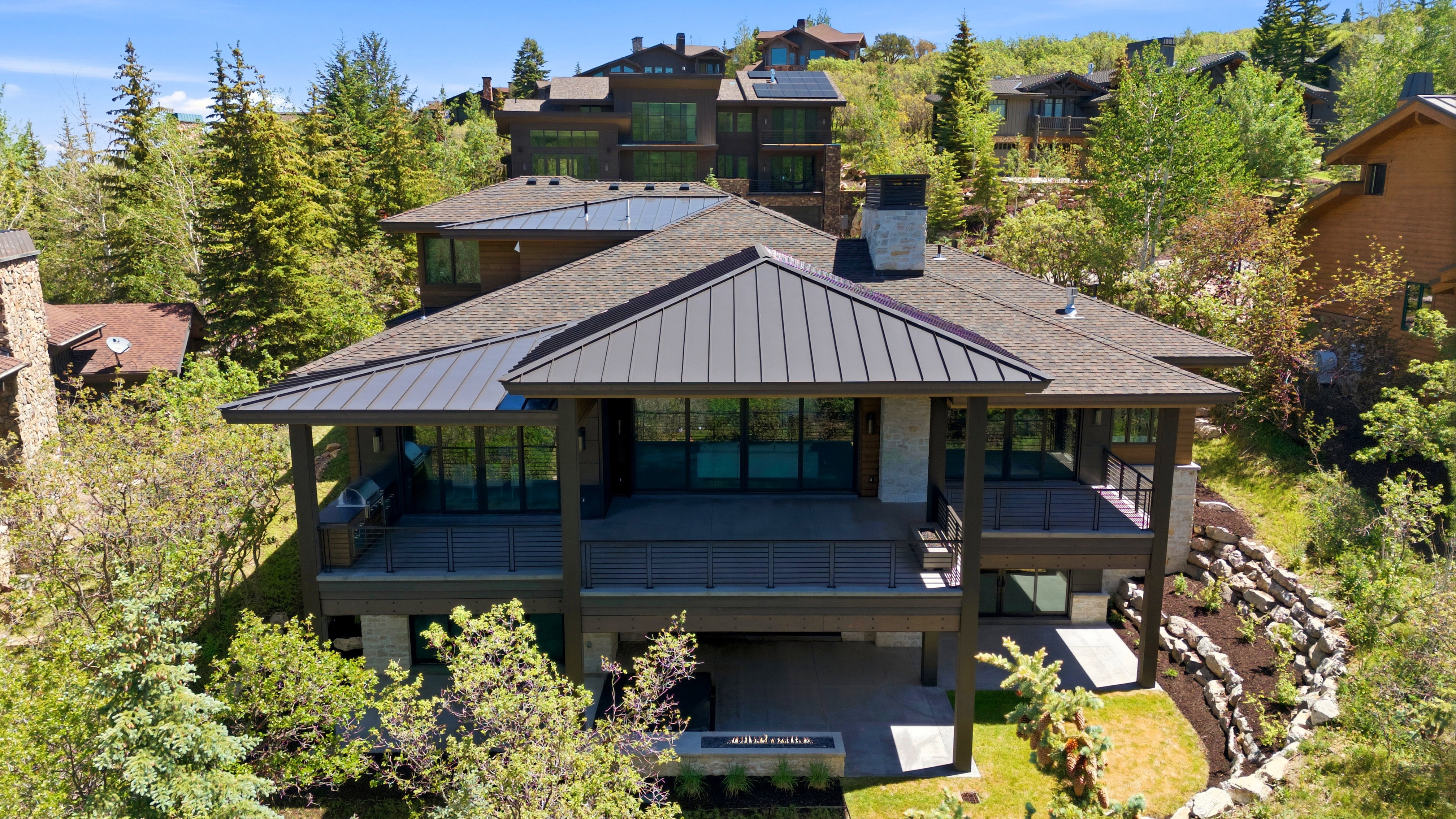 Contemporary home exterior featuring a standing seam metal roof, stone chimney, expansive outdoor deck with glass railings, and a lower-level patio with a built-in fire pit