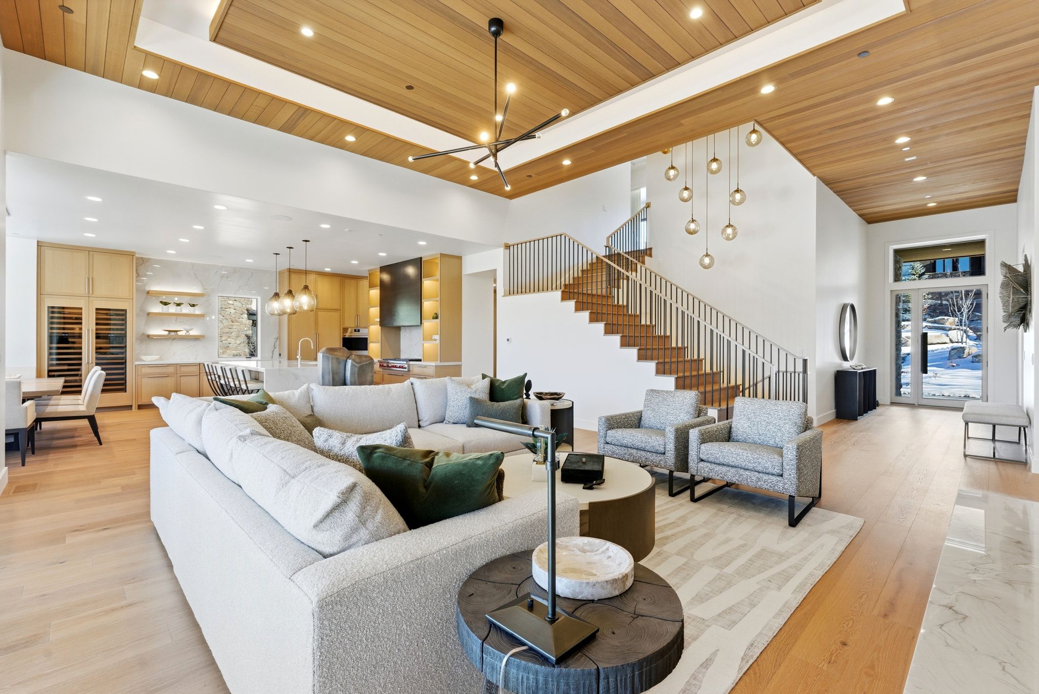 Open-concept living area featuring light wood-finish flooring, a two-story ceiling with recessed lighting and wood panel accents, and a built-in kitchen with integrated appliances