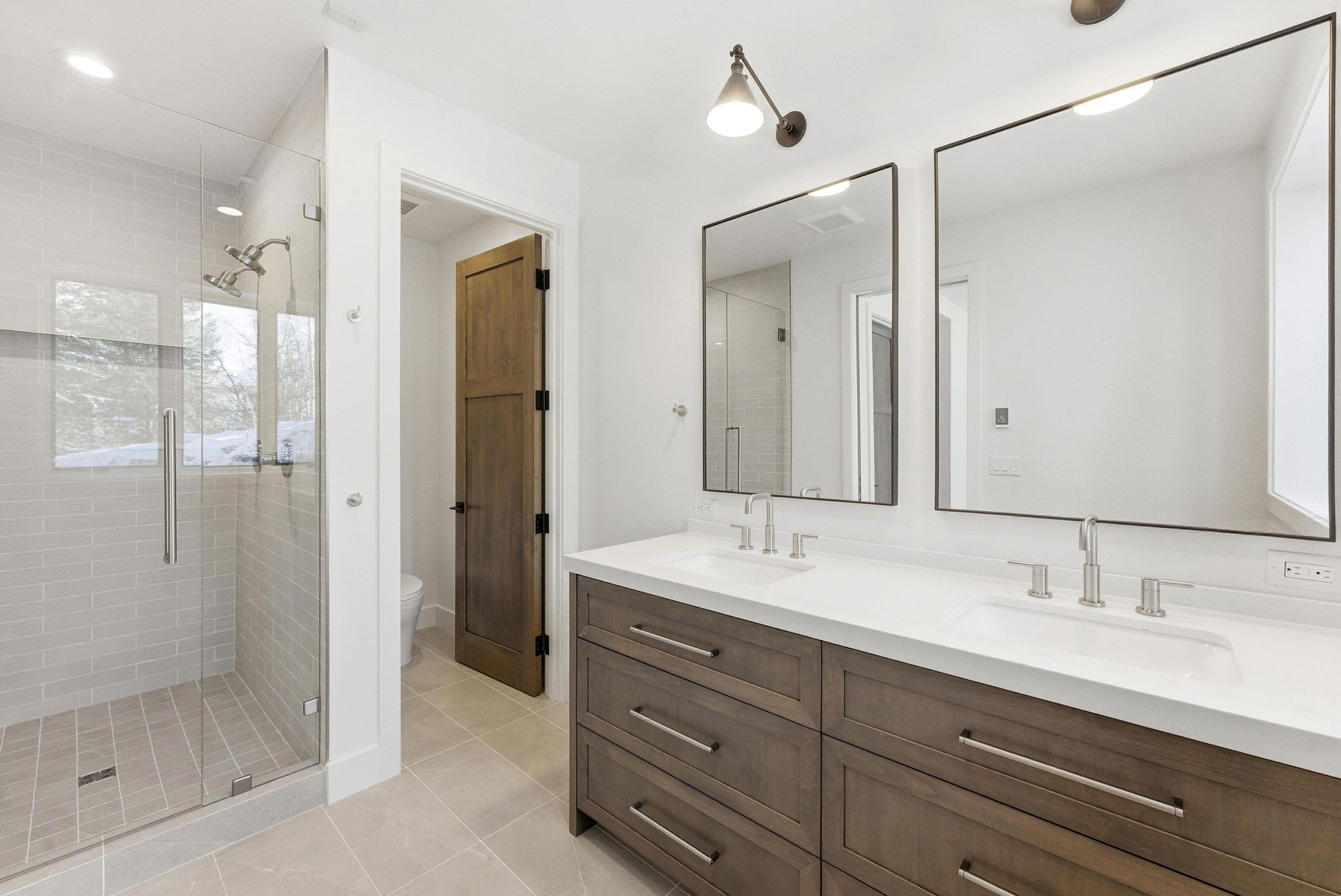 Bathroom featuring a glass-enclosed shower with subway tile surround, a dual vanity with a white countertop and wood-finish cabinetry, two rectangular framed mirrors, and light tile flooring