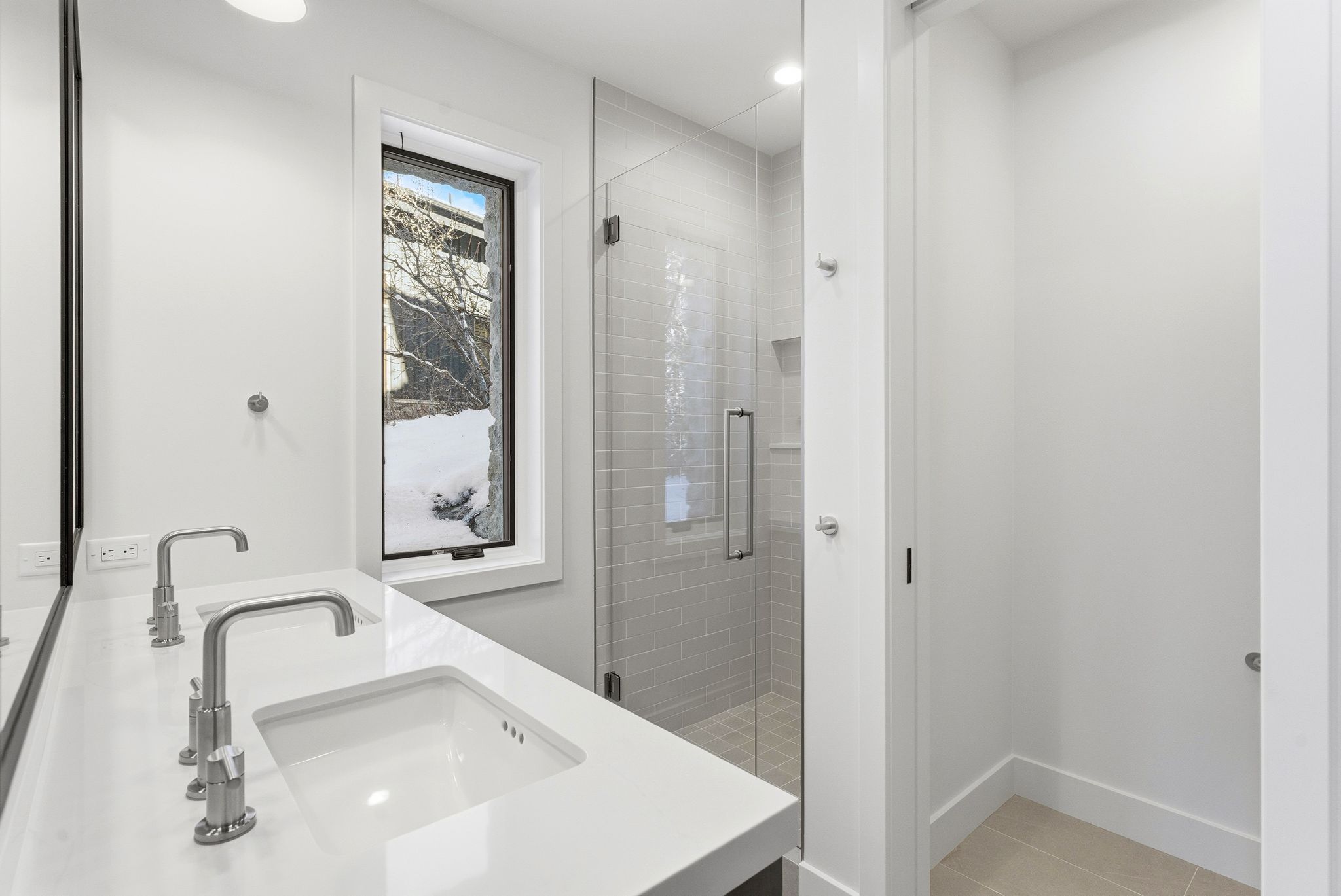 Bathroom featuring a dual vanity with integrated sinks and wall-mounted faucets, a frameless glass shower enclosure with subway tile surround, a tall vertical window, and light-toned flooring