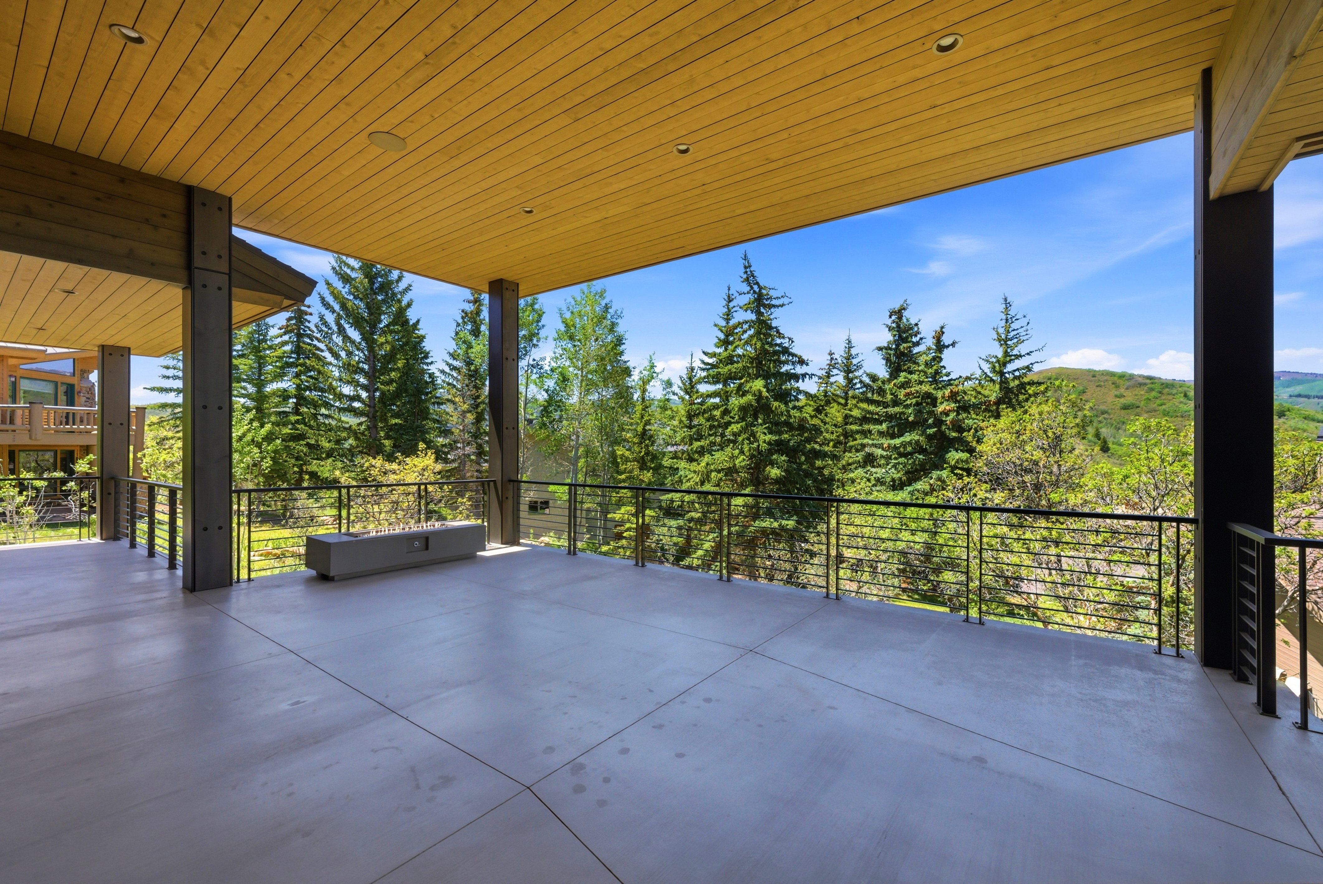 Expansive concrete patio featuring a wood-panel ceiling with recessed lighting, metal support columns, a linear built-in fire pit, and a cable railing system