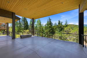 Expansive concrete patio featuring a wood-panel ceiling with recessed lighting, metal support columns, a linear built-in fire pit, and a cable railing system