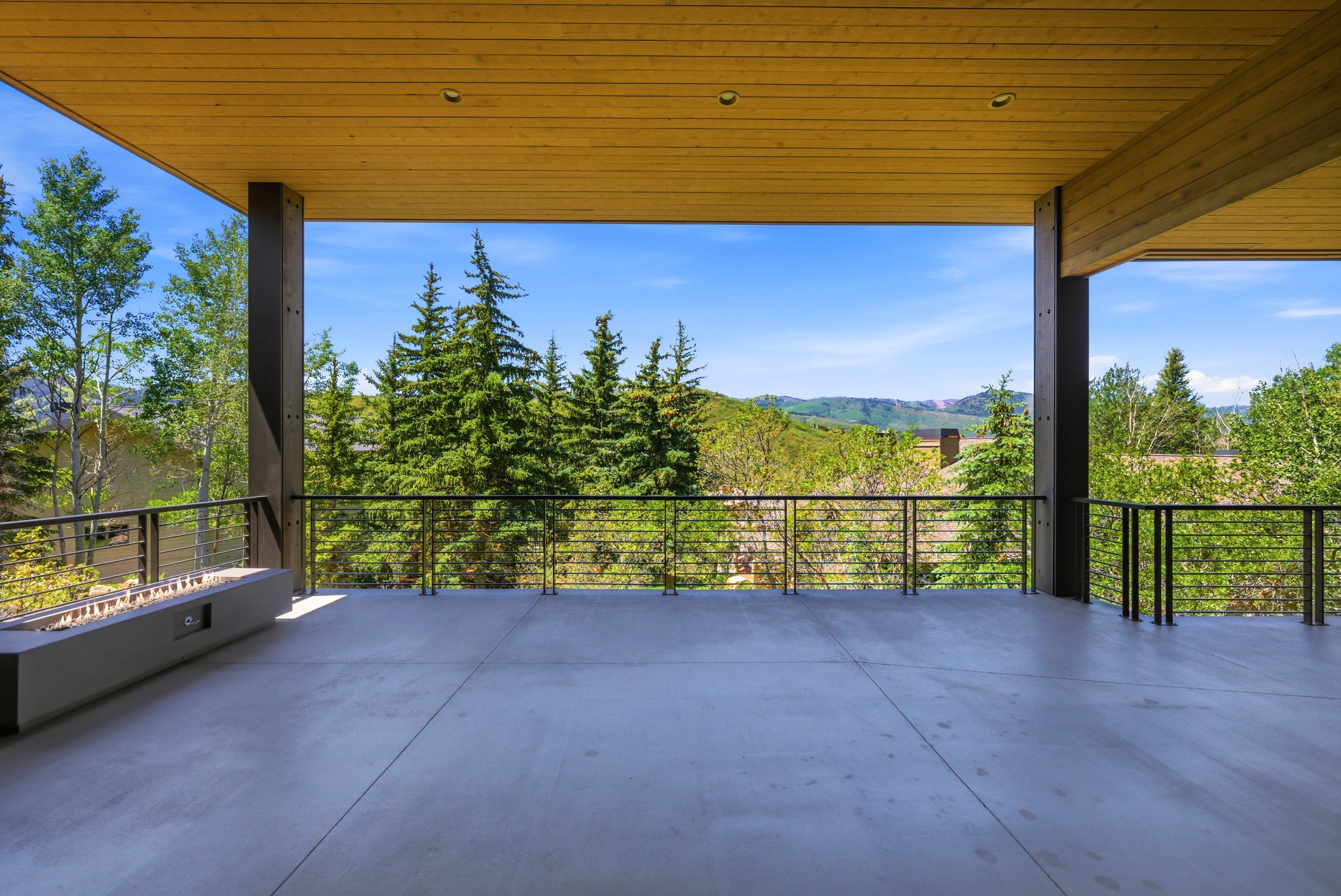Spacious covered outdoor deck featuring a concrete floor, wood-paneled ceiling, and a built-in linear fire pit