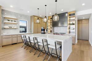 Expansive kitchen featuring a large central island with seating, light wood-finish cabinetry, integrated appliances, and a full-height stone-look backsplash