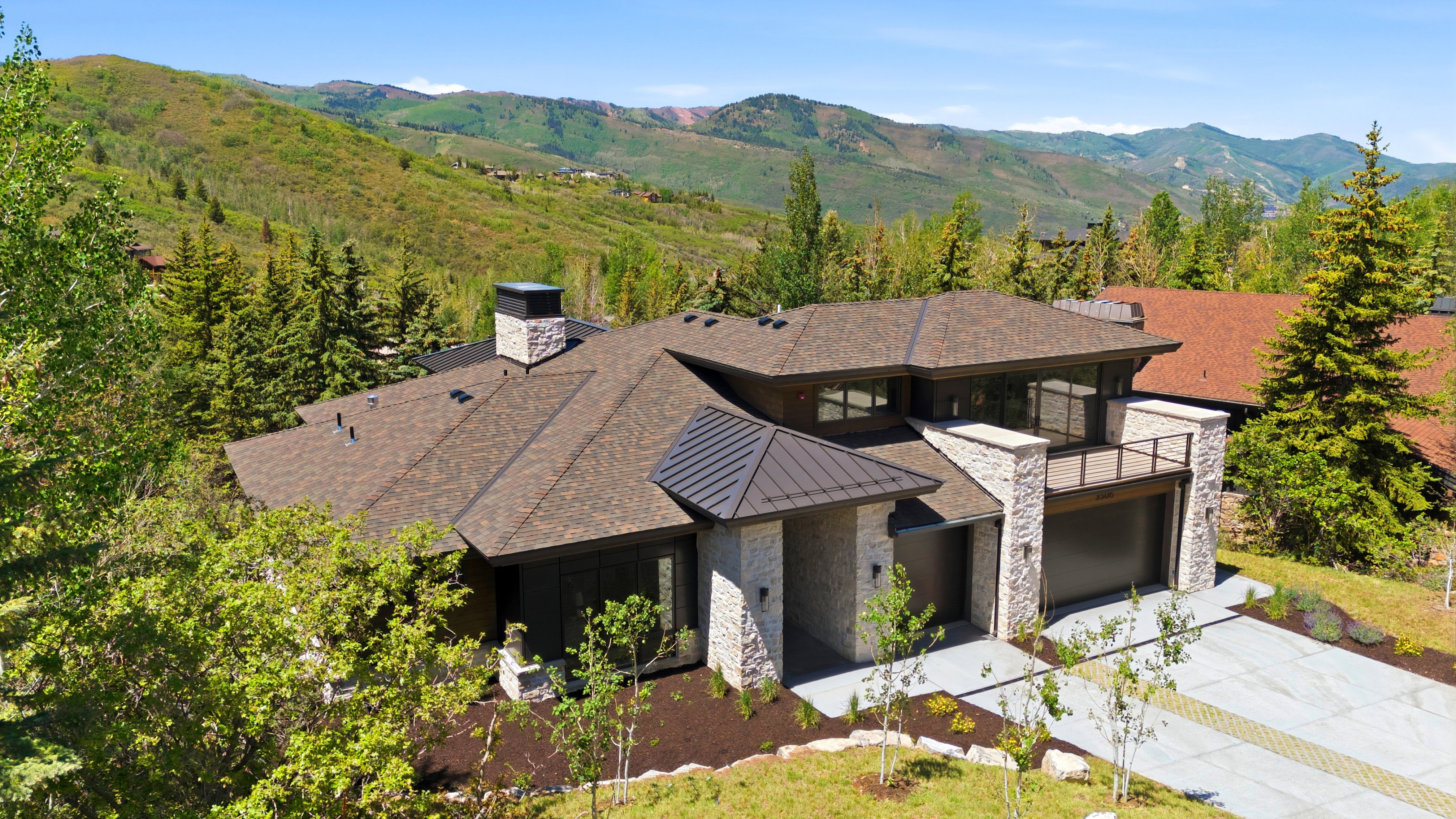 Contemporary mountain residence featuring a multi-gabled roofline, stone and wood-tone exterior accents, a prominent balcony with metal railing, an integrated garage, and a concrete driveway