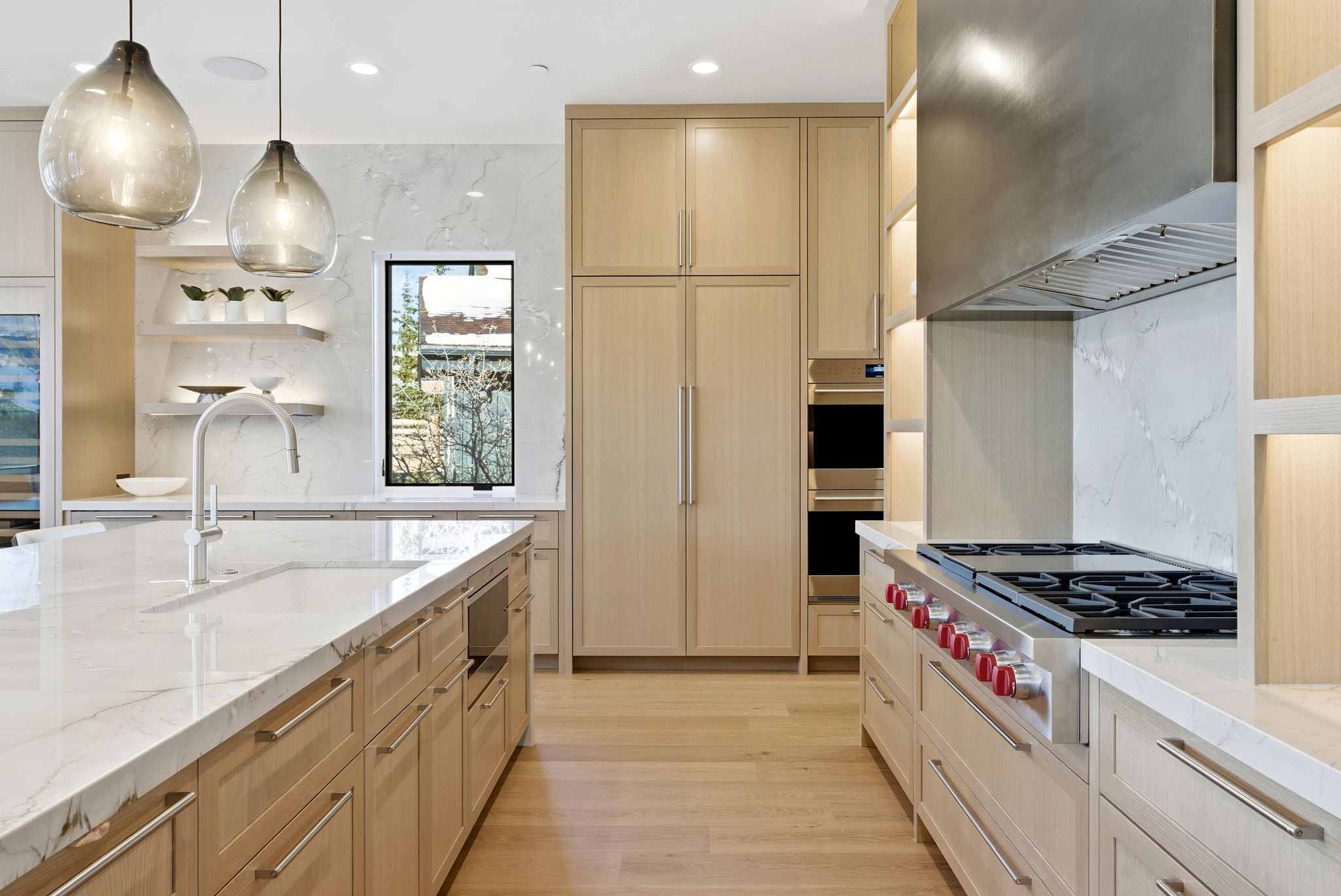 Gourmet kitchen featuring a large island with an integrated sink and gooseneck faucet, light wood-finish cabinetry, white countertops, built-in stainless steel appliances, and a gas range with an overhead exhaust hood