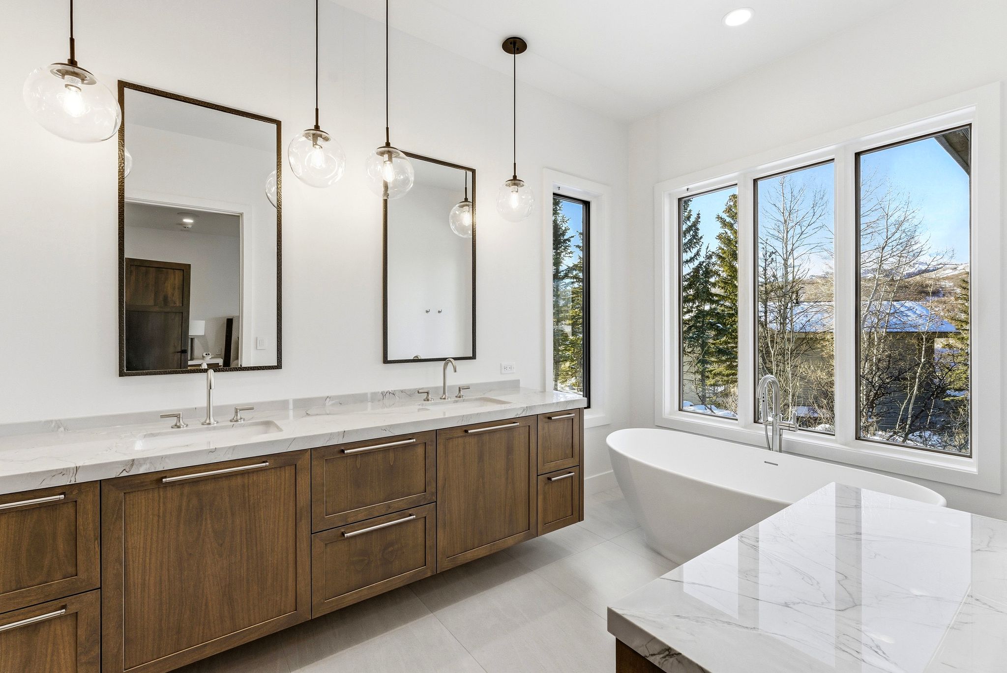 Modern bathroom featuring a freestanding tub, large windows, wood-finish double vanity with stone countertops, and pendant lighting