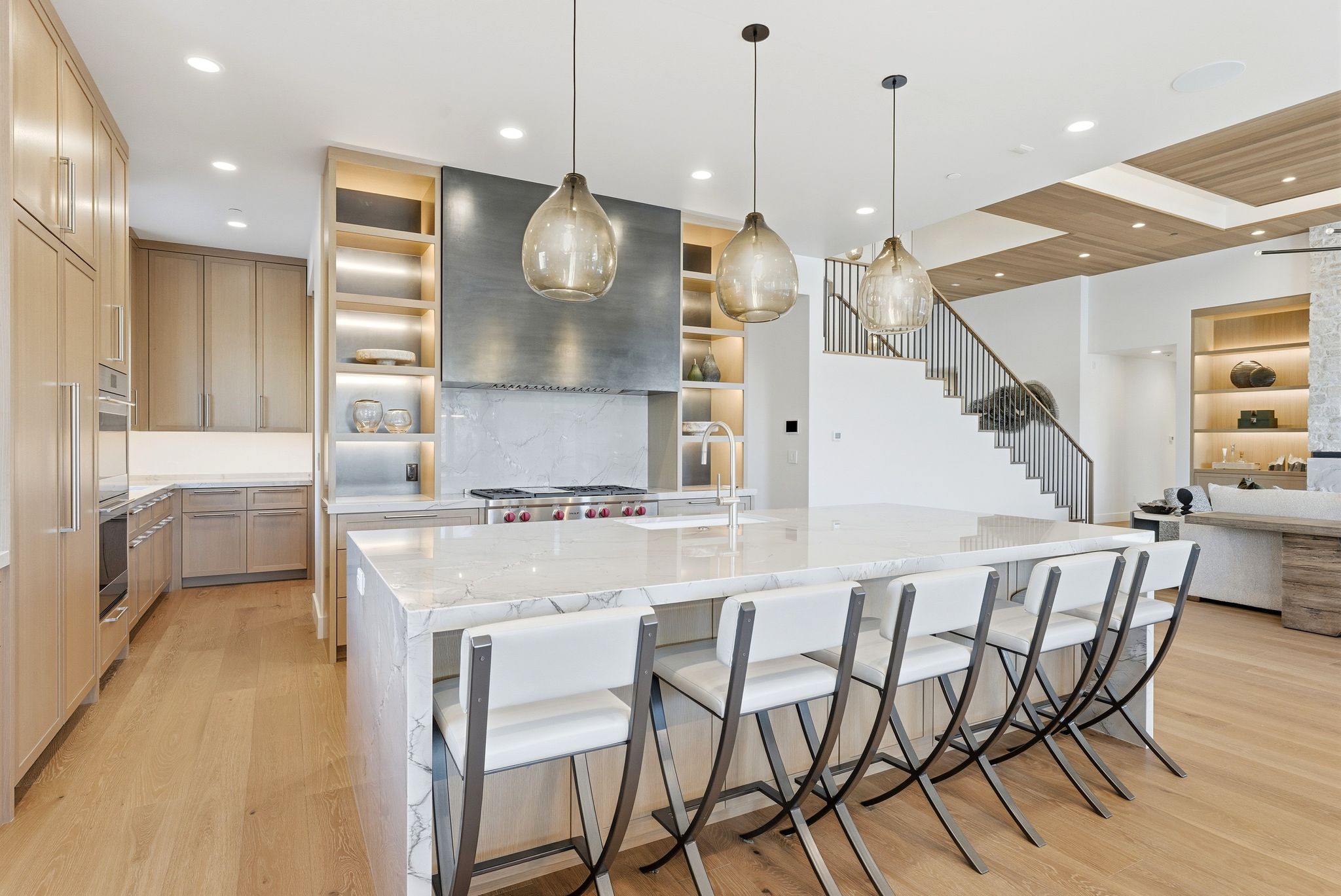 Gourmet kitchen featuring a waterfall island with seating, pendant lighting, professional-grade range with a custom hood, light wood-finish cabinetry, and built-in shelving