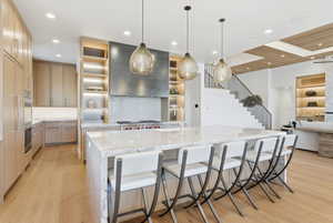 Gourmet kitchen featuring a waterfall island with seating, pendant lighting, professional-grade range with a custom hood, light wood-finish cabinetry, and built-in shelving