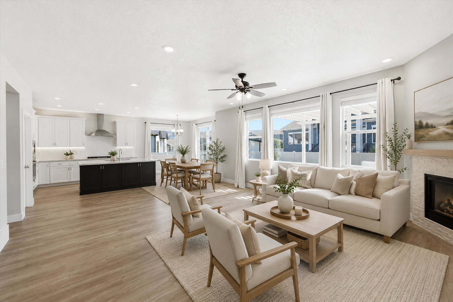 Open-concept living space featuring wood-finish flooring, a contemporary kitchen with white cabinetry and a dark island, a dining area with a chandelier, a living area with large windows, and a stone-clad fireplace