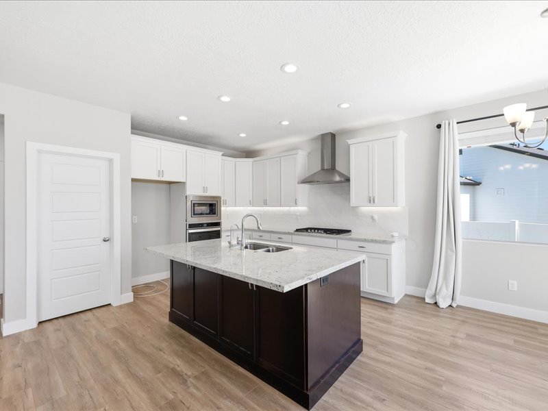 Kitchen featuring a dark-toned island with a light-toned countertop, white cabinetry, stainless steel range hood, built-in stainless steel oven and microwave, and wood-finish flooring