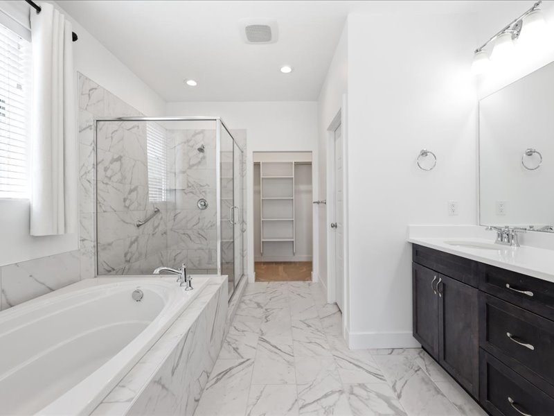 Bathroom featuring a built-in tub, a glass-enclosed shower with a grab bar, a single vanity with a white countertop and dark cabinetry, and white tile flooring with gray veining