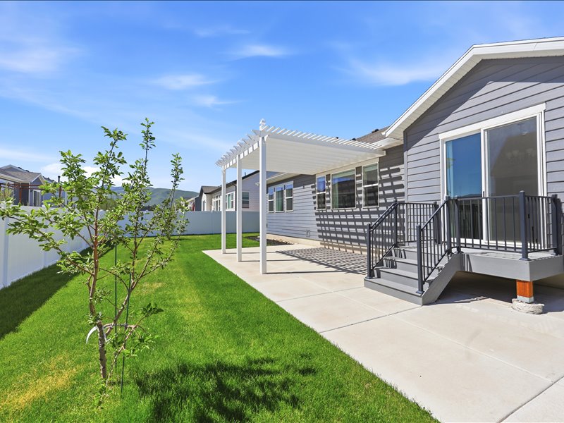 Backyard featuring a concrete patio with a white pergola, leading to a raised deck with dark railings