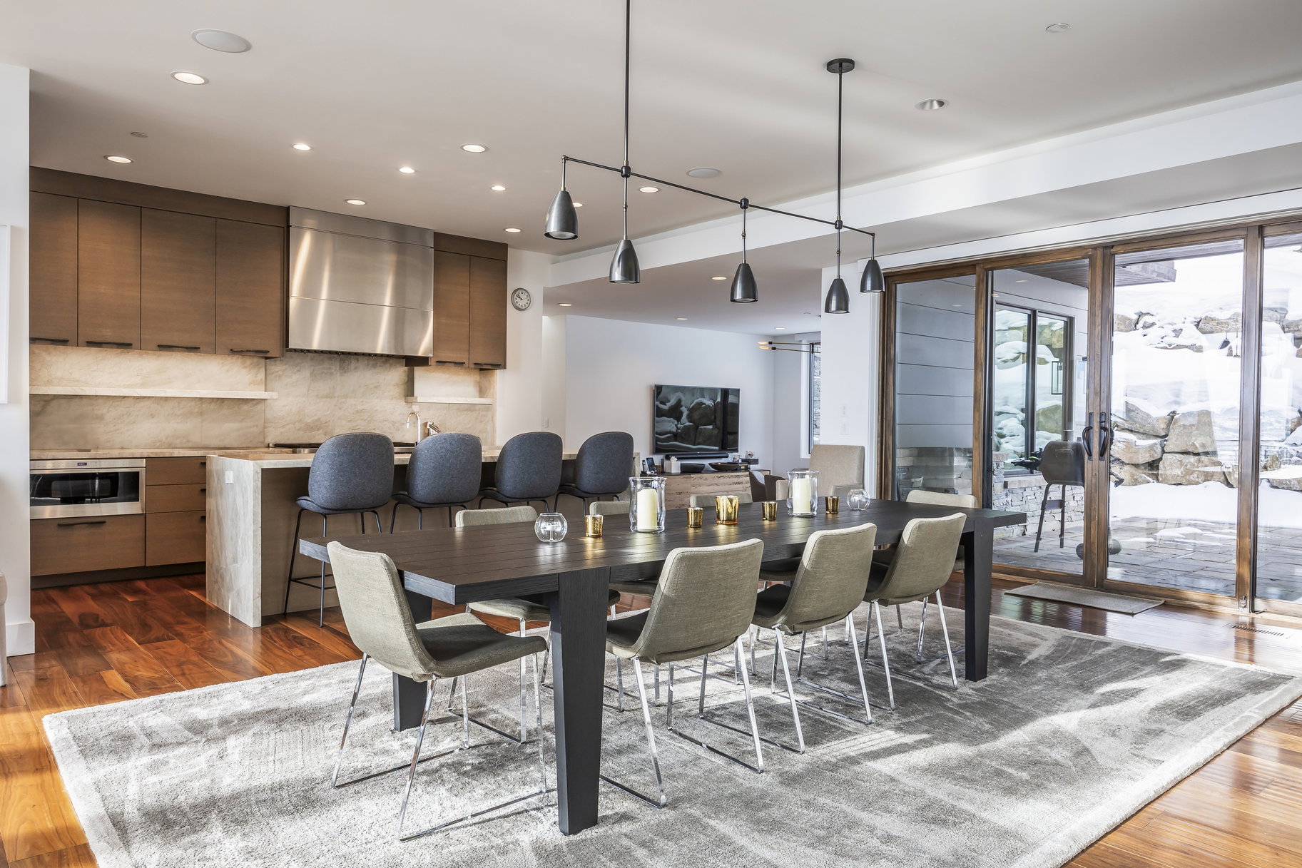 Open-concept living area featuring rich wood-finish flooring, a modern kitchen with integrated appliances, a spacious island with seating, a large dining area, and expansive sliding glass doors