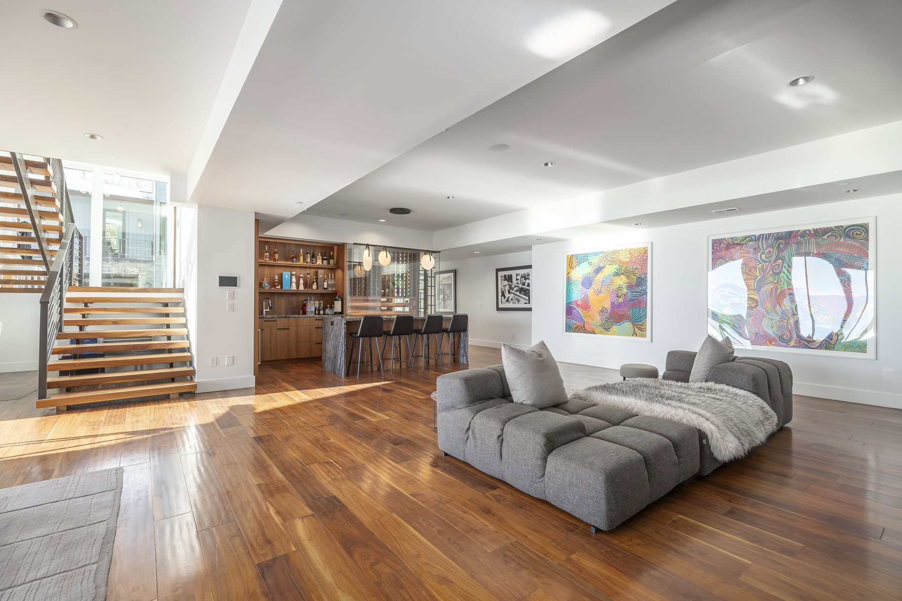 Spacious open-concept interior featuring hardwood flooring, a built-in wet bar with seating, a contemporary staircase with metal railings, recessed lighting, and expansive wall space for art