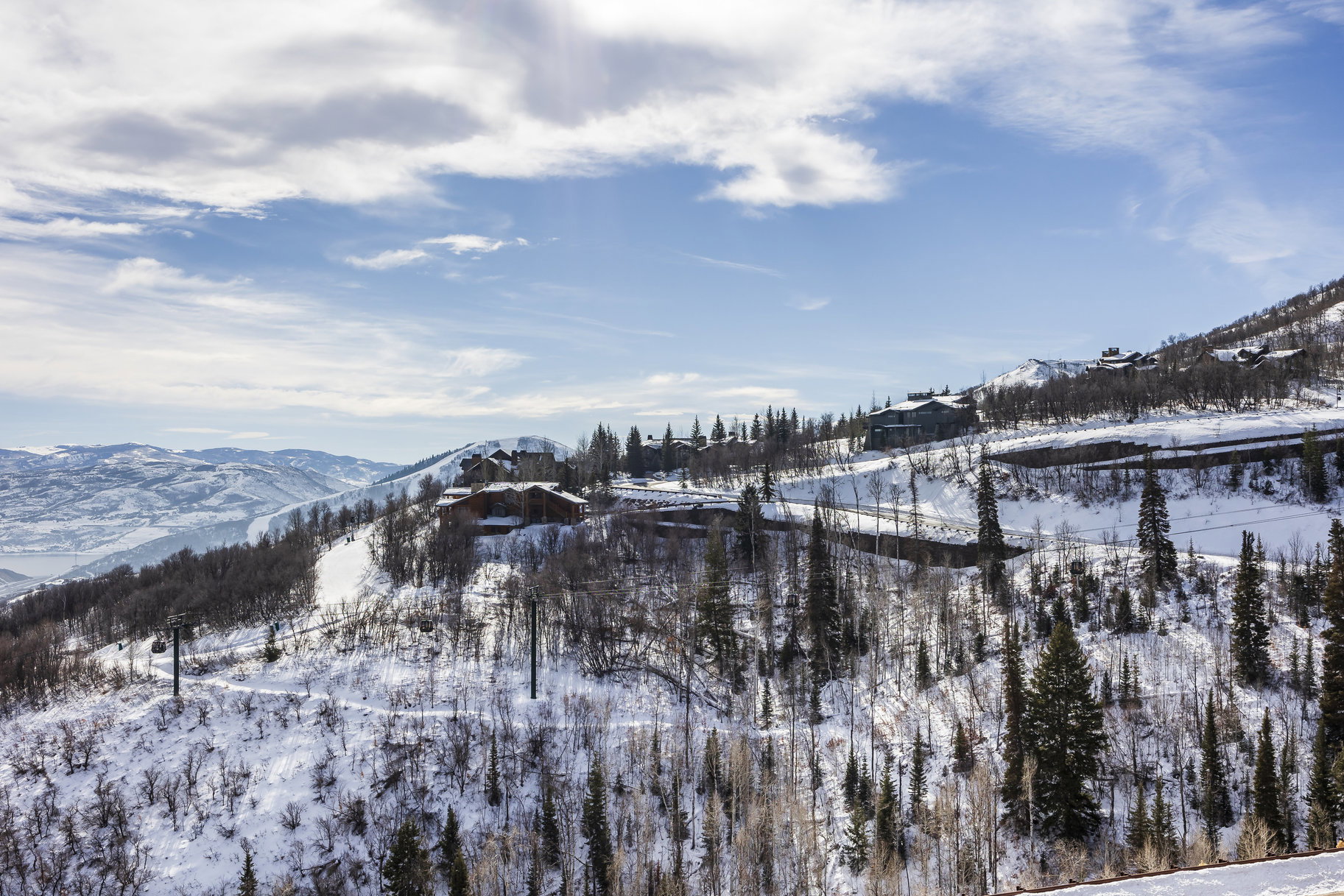 Snow-covered mountain landscape featuring multiple properties, dense evergreen and deciduous trees, and a distant body of water